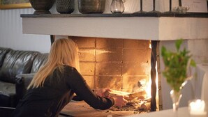 Une personne assise près d’une cheminée où le feu brûle, avec un vase contenant une bougie, et un canapé en cuir noir en arrière-plan.