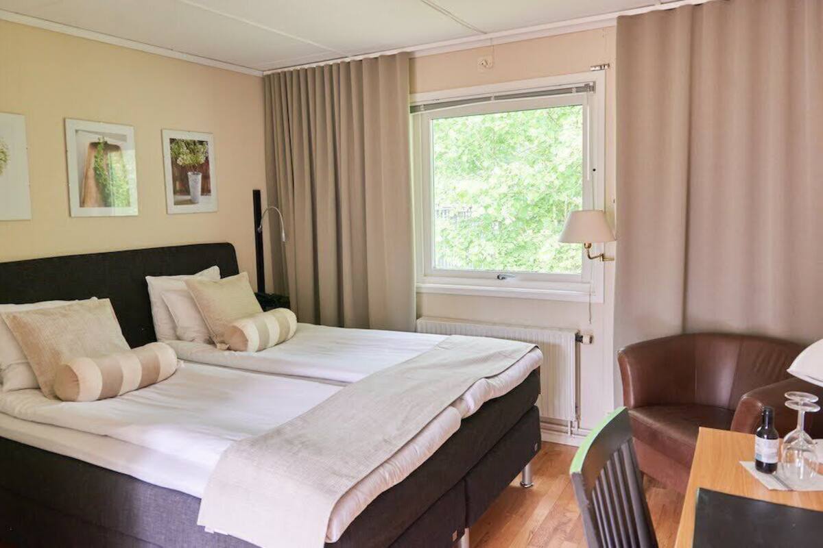 Une chambre d’hôtel avec un lit, une fenêtre avec des rideaux, une chaise et une table sur laquelle se trouvent une bouteille et un verre.