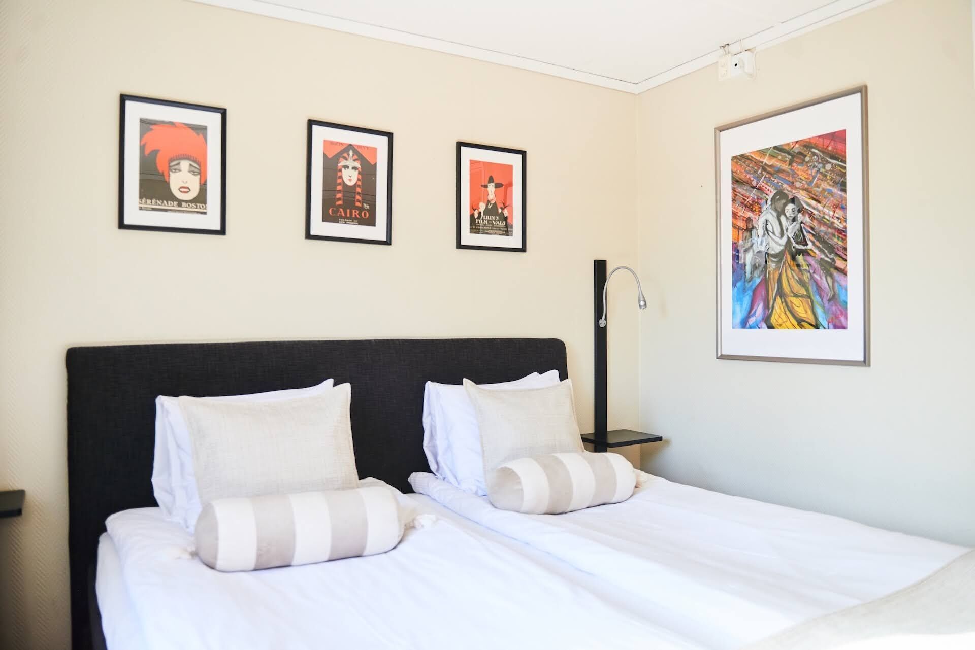 Habitación de hotel con una cama, dos almohadas, una mesita de noche y tres carteles enmarcados en la pared.