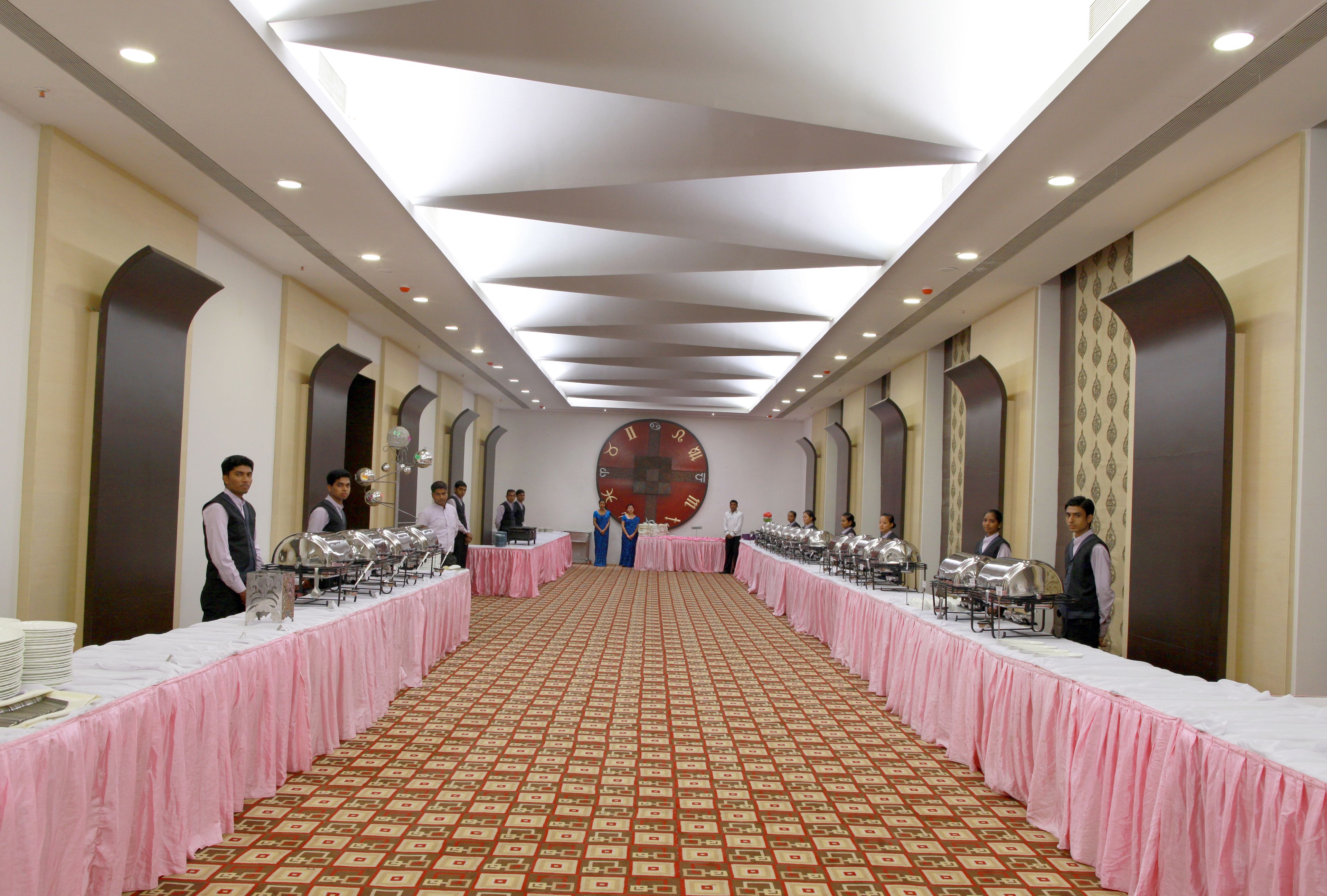 A banquet hall with a long table covered in a pink tablecloth, a patterned carpet, and a large clock on the wall.