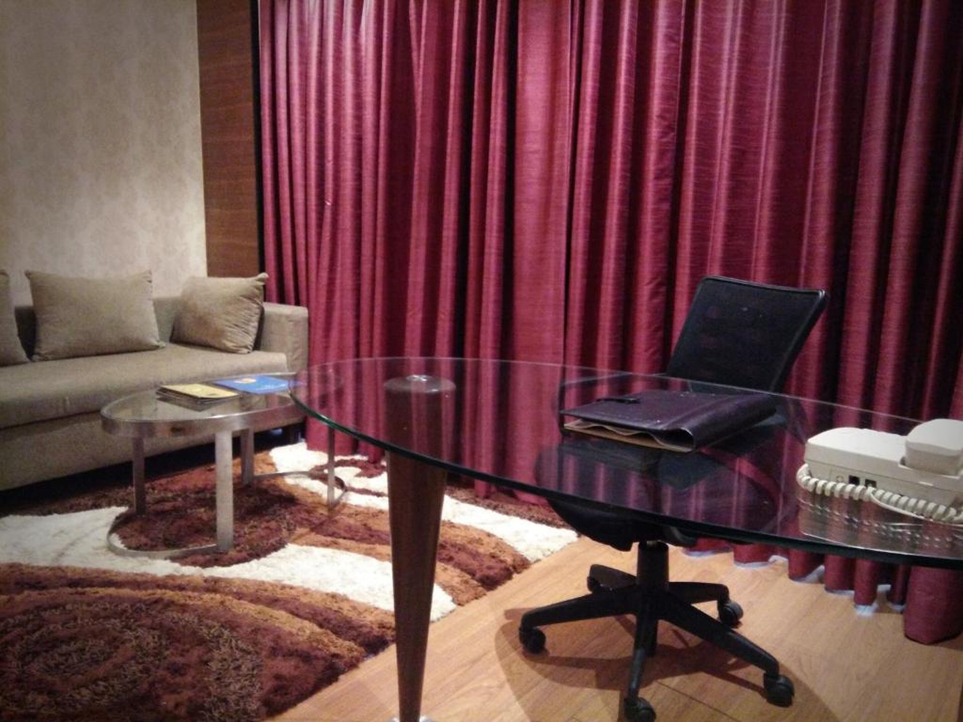 A modern living room with a glass table, a black office chair, a sofa, and a red curtain backdrop.