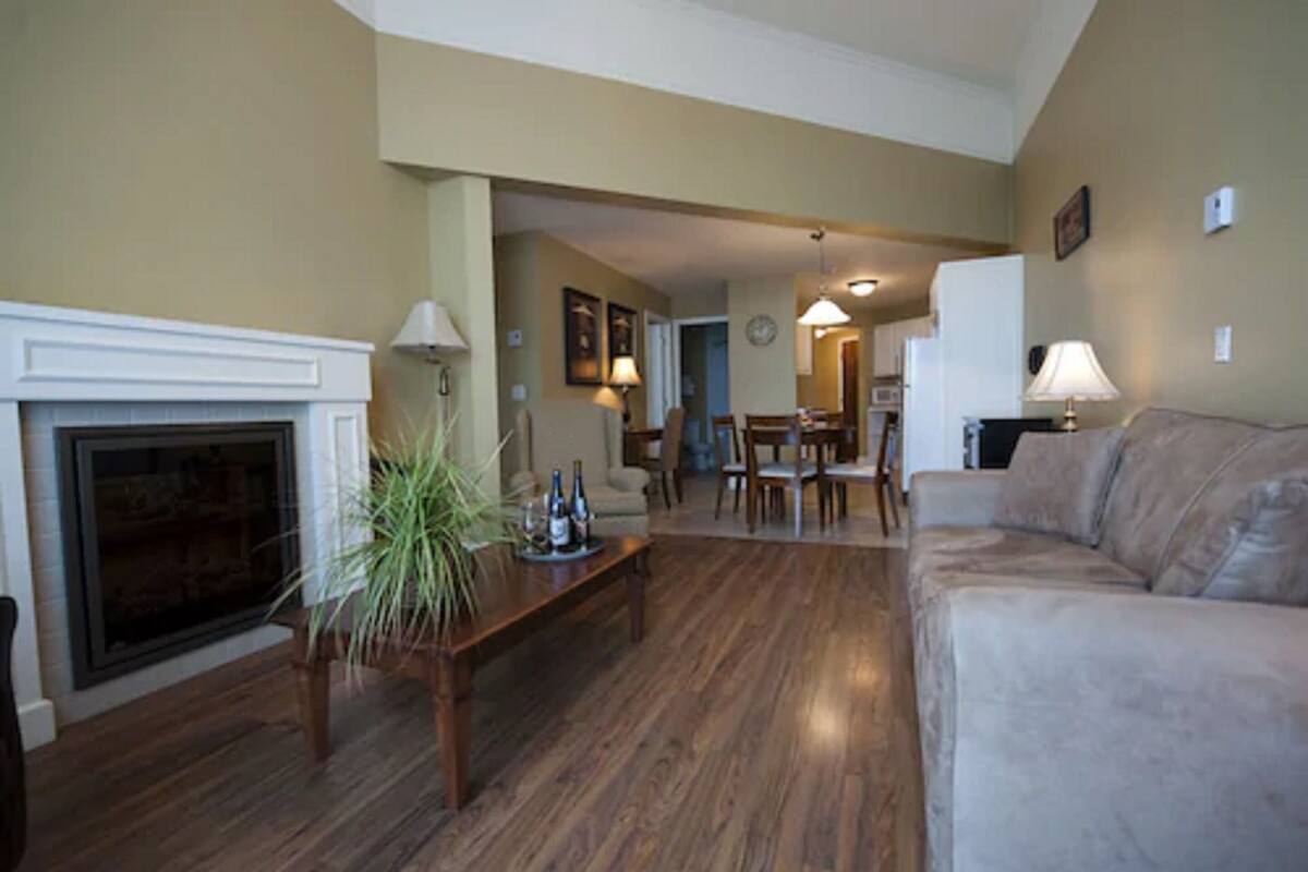 A living room with a fireplace, a sofa, a dining area, and a kitchen in the background.