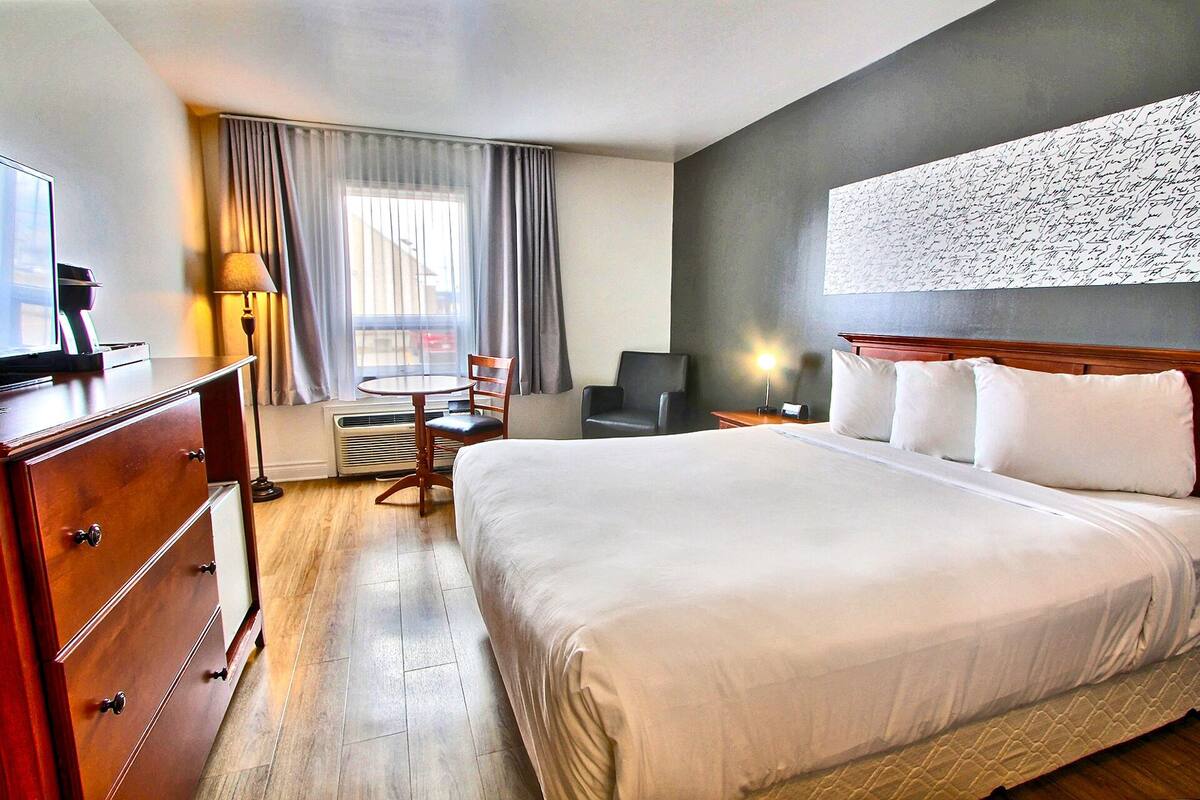 A hotel room with a large bed, a desk with a computer, a chair, a window with curtains, and a wall-mounted artwork.