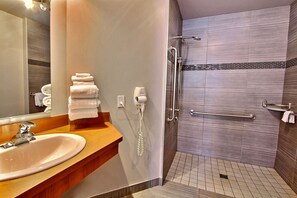 A bathroom with a sink, mirror, towel rack, and a walk-in shower with grab bars. - Hôtel L'Empress (Rimouski)