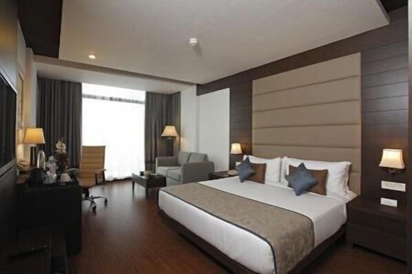 A hotel room with a large bed, two bedside tables, a desk, a chair, and a window with curtains. - SK Premium Park (New Delhi)