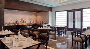 A dining area with tables set for breakfast, a buffet line, and a large window with blinds. - SK Premium Park (New Delhi)