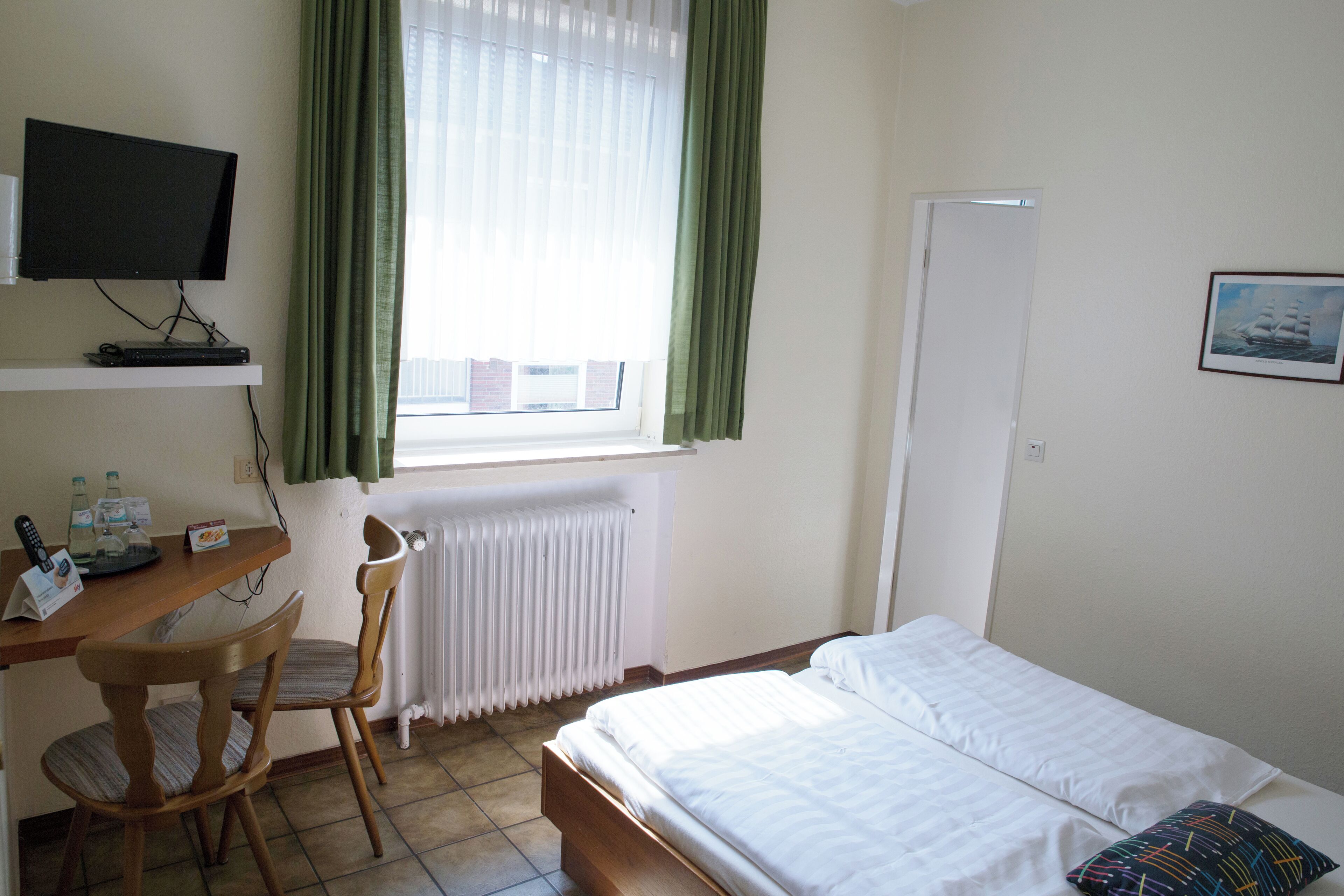 A hotel room with a bed, a desk with a computer, a chair, a window with curtains, and a radiator.