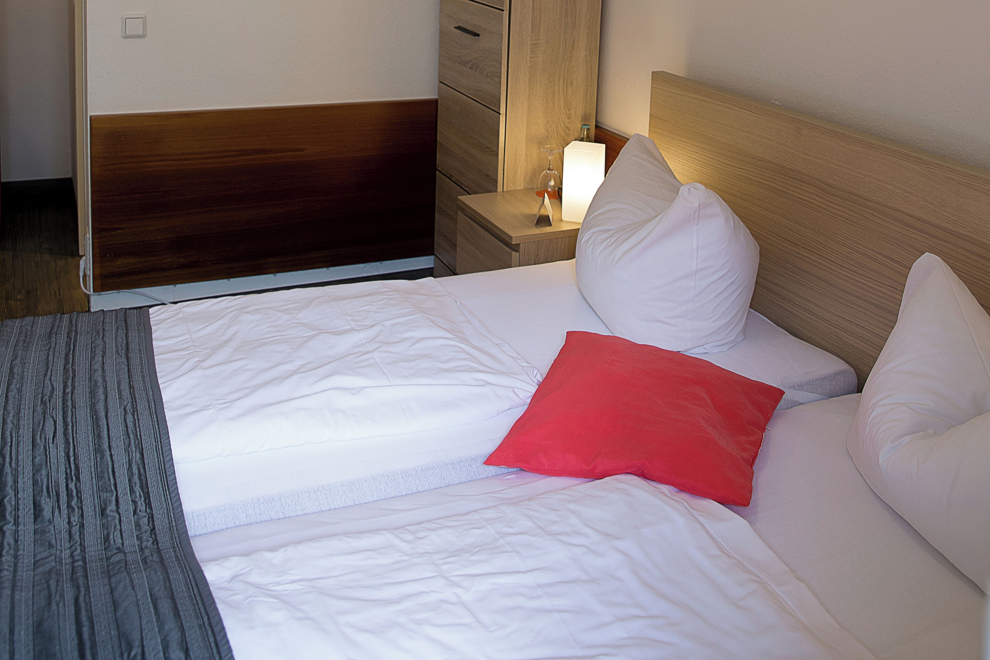A neatly made bed with white bedding and a single red pillow, a wooden headboard, and a bedside table with a lamp.