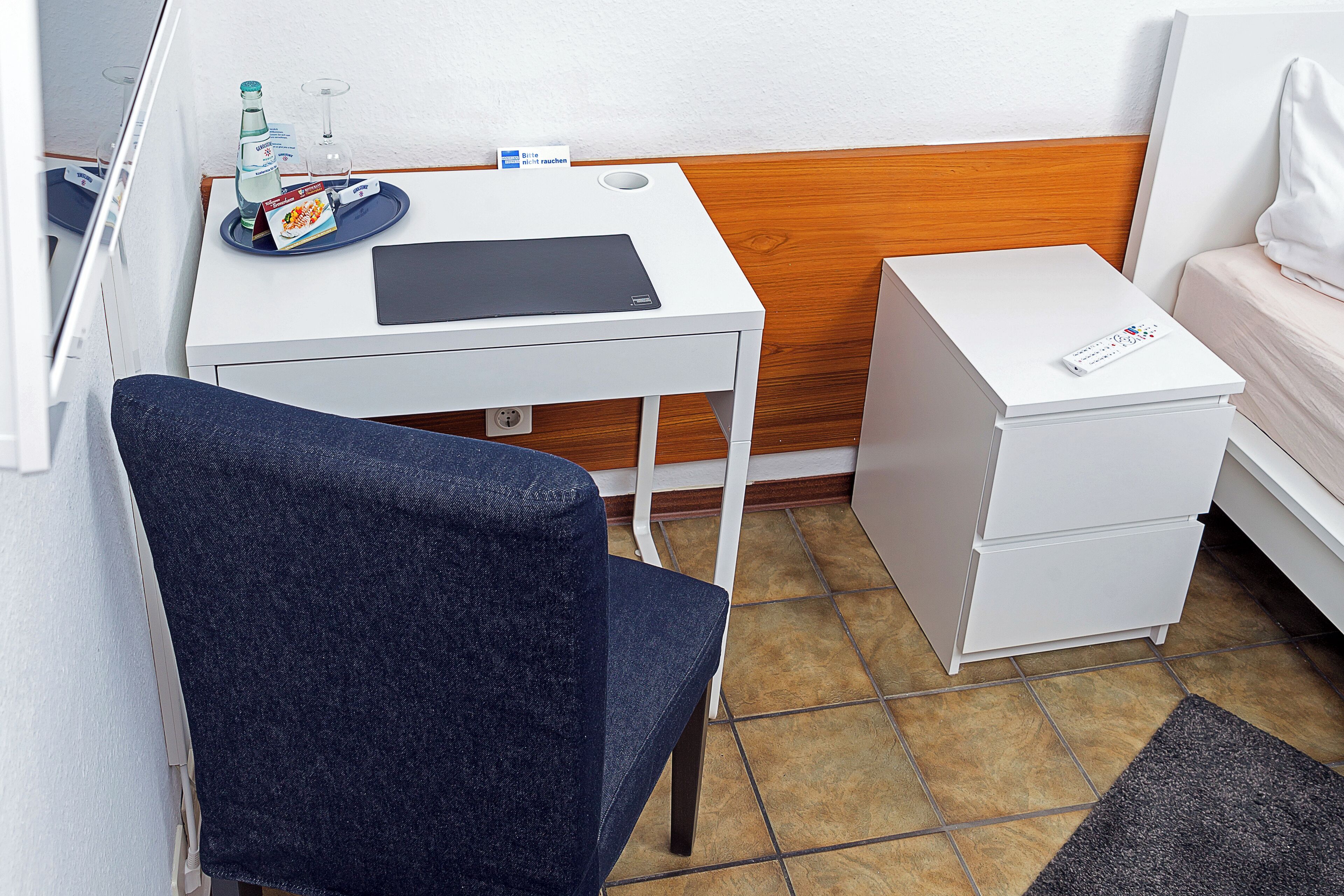 A small, compact room with a white desk, a chair, a bedside table, and a bed.