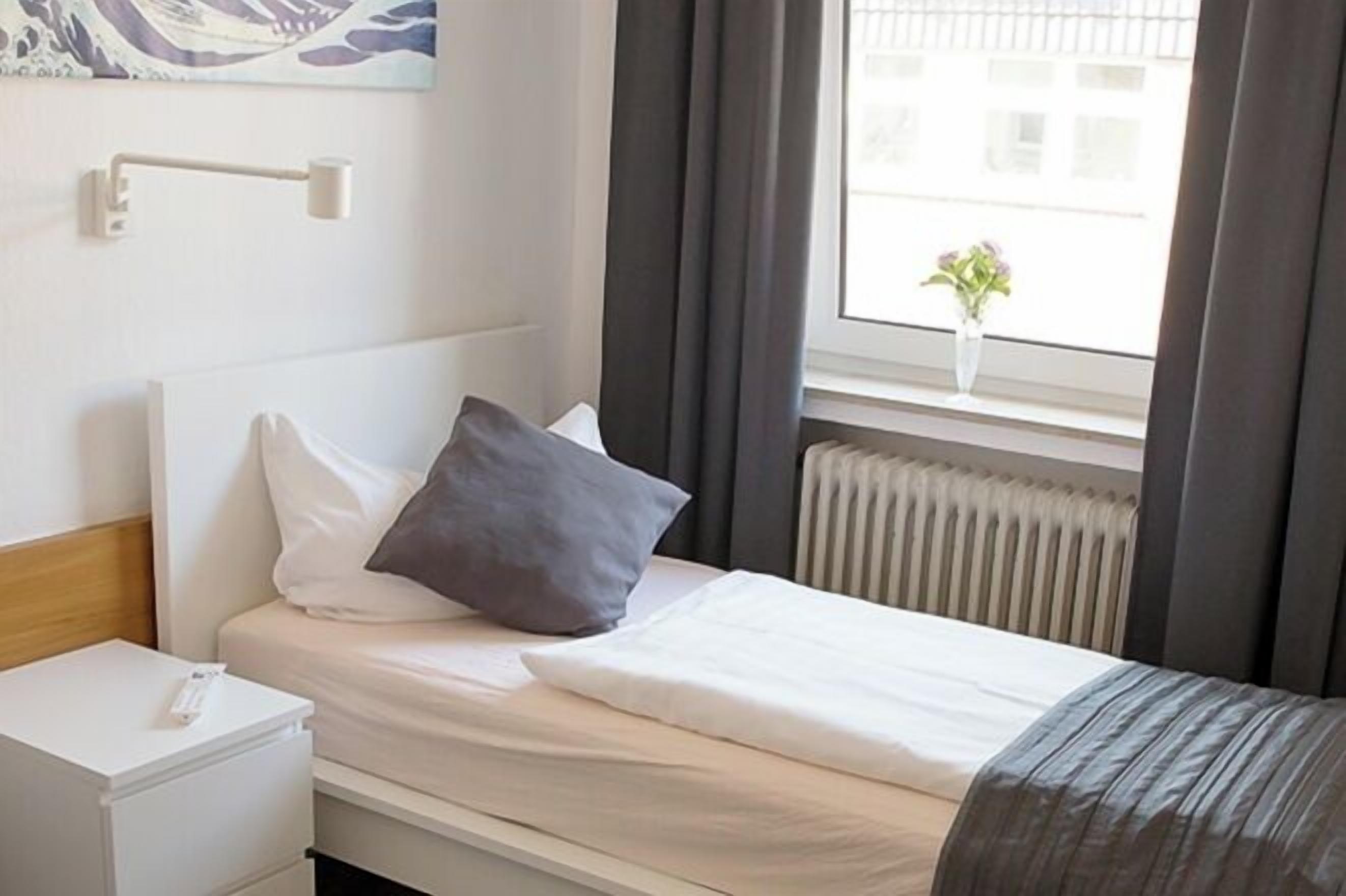 A neatly made bed with white and gray bedding, a white bedside table with a lamp, and a window with a view of buildings and a plant.
