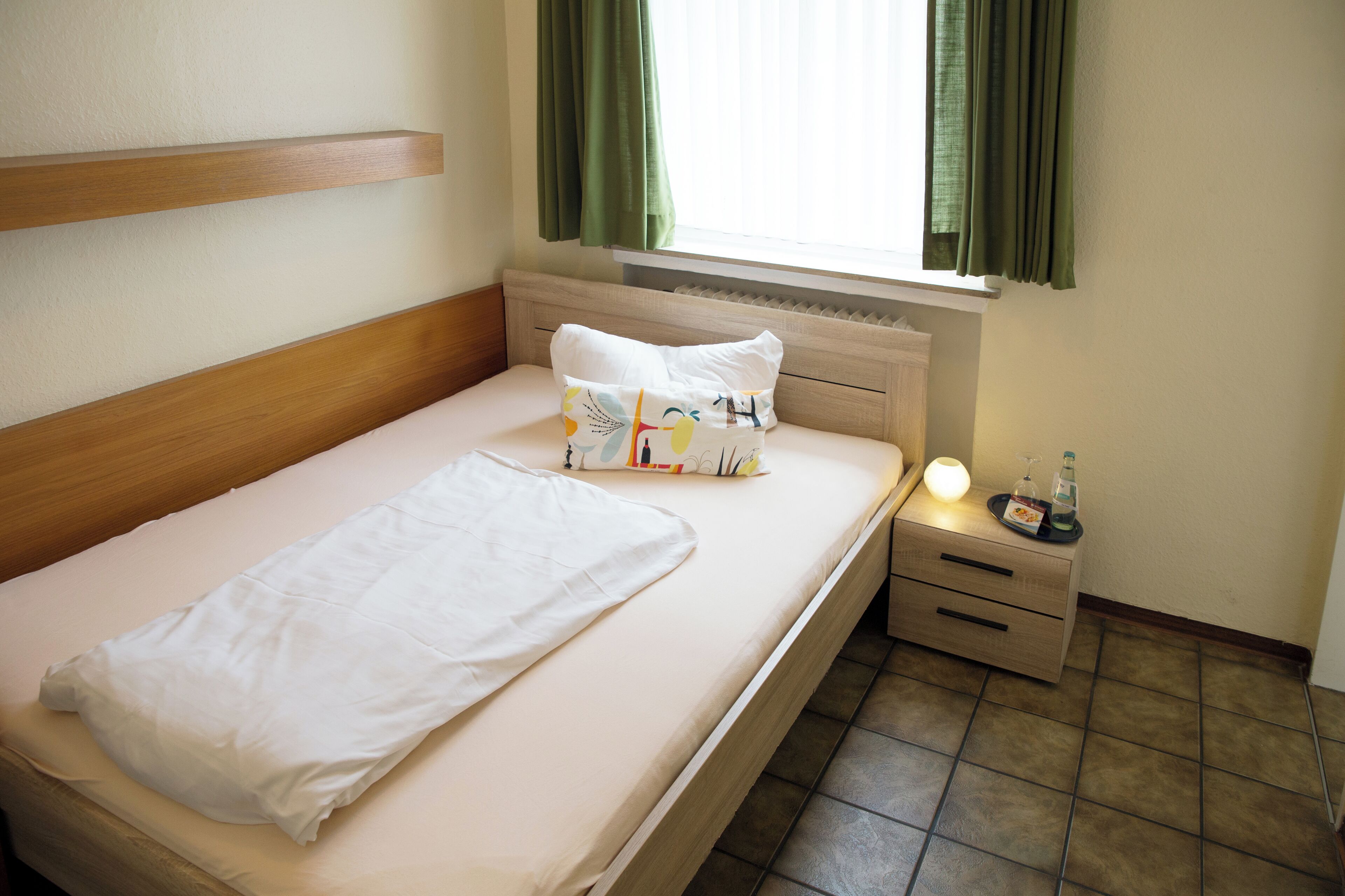 A single bed with a wooden headboard, a bedside table with a lamp, and a window with curtains.