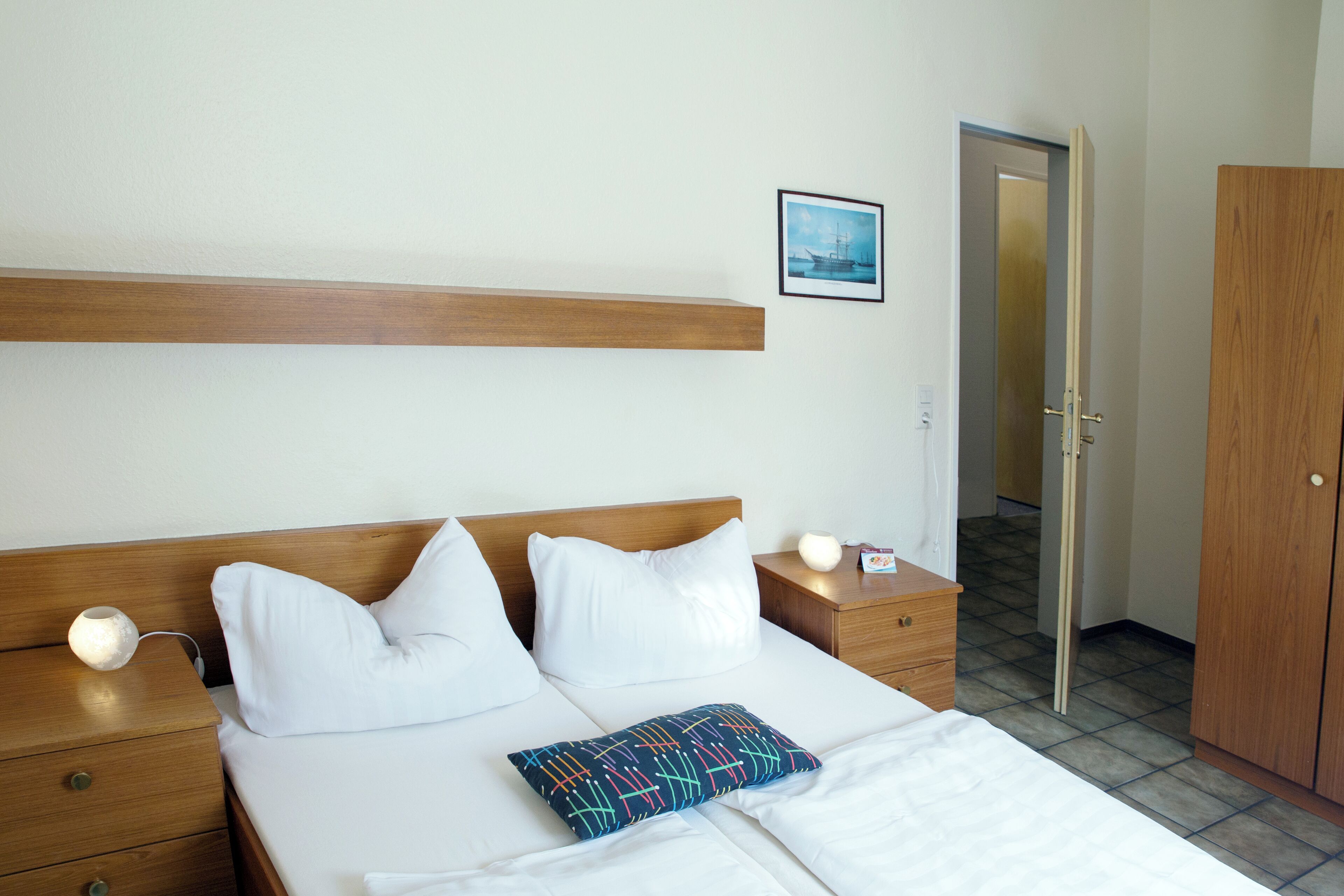 Hotellihuone, jossa on puinen sängynpääty, valkoisilla lakanoiden peitetty sänky, värikäs tyyny, yöpöytälamput, yöpöytä ja vaatekaappi.