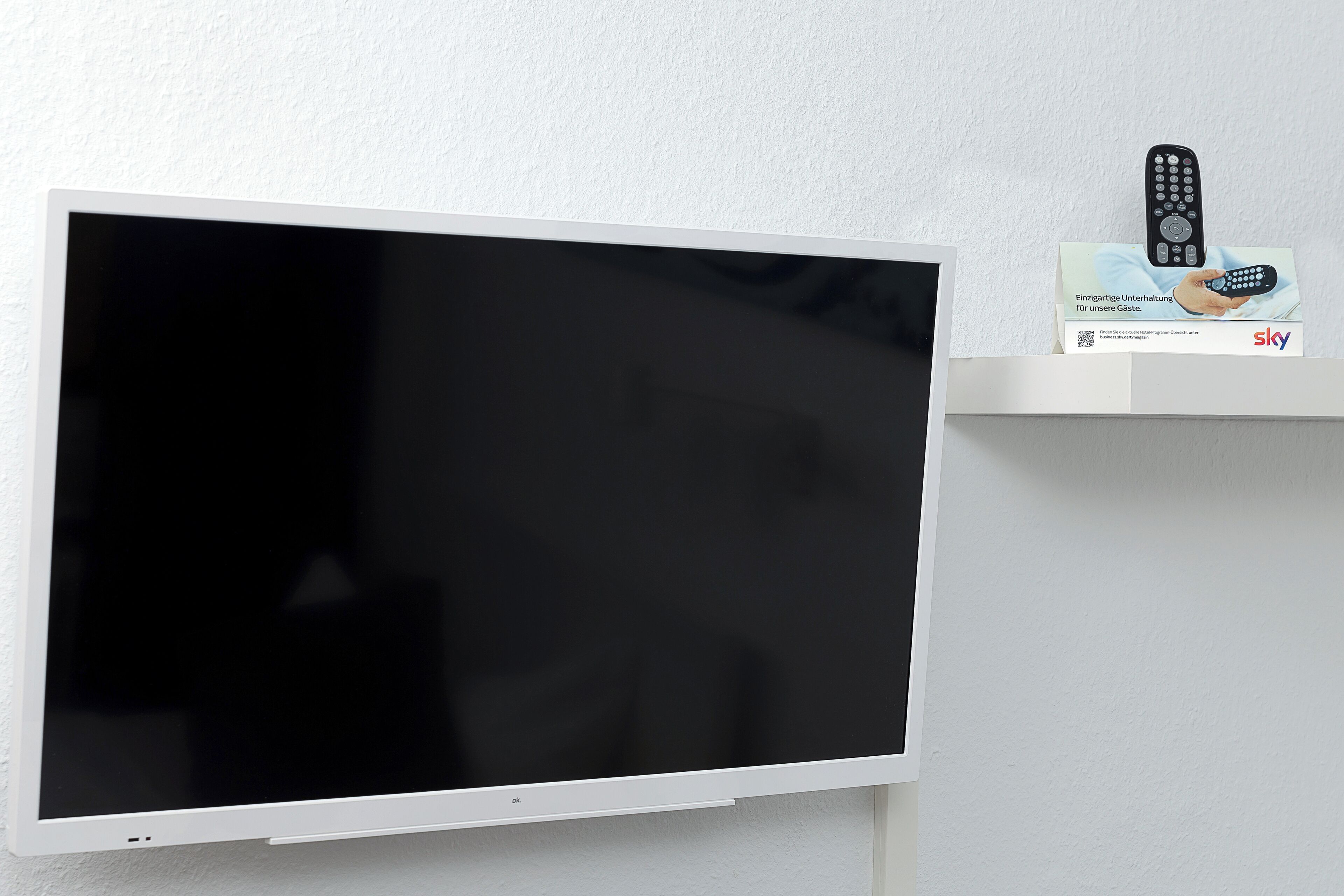 A flat-screen TV mounted on a wall, a remote control on a shelf, and a box with a remote control image.