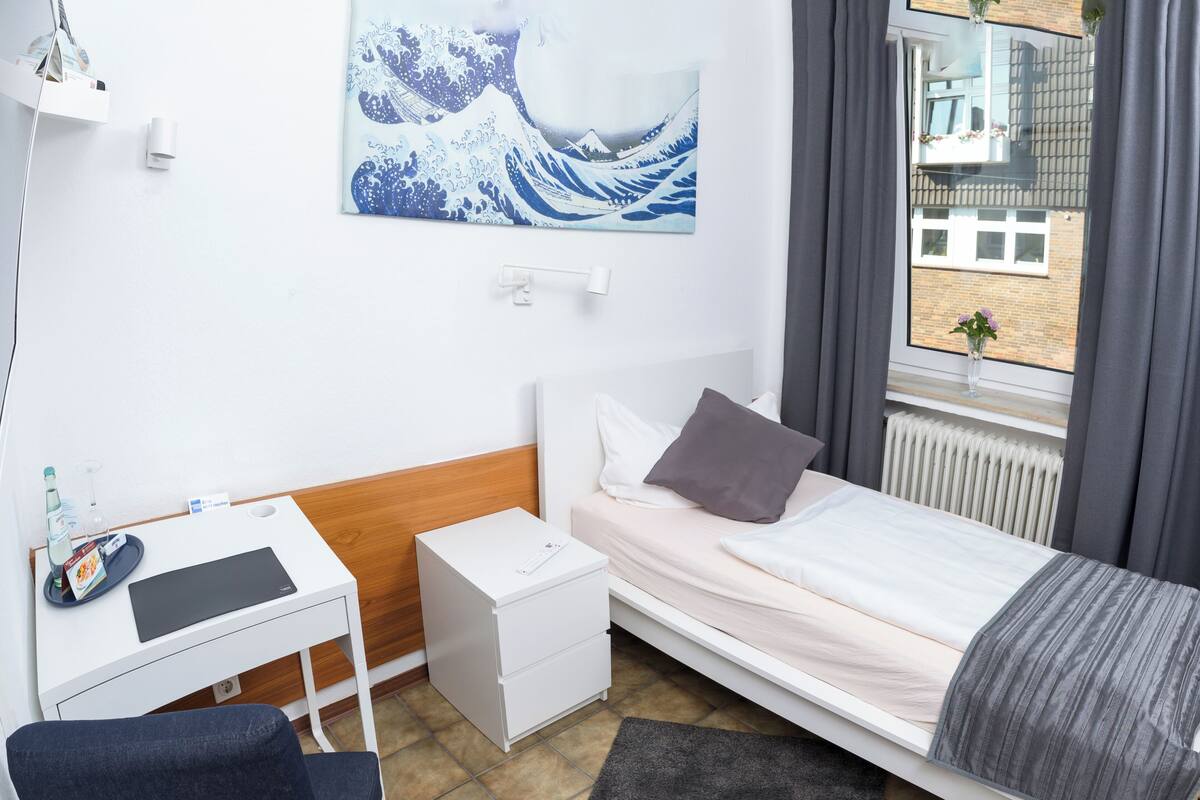 A hotel room with a bed, a desk, a chair, a nightstand, a window with a view of a building, and a painting on the wall.