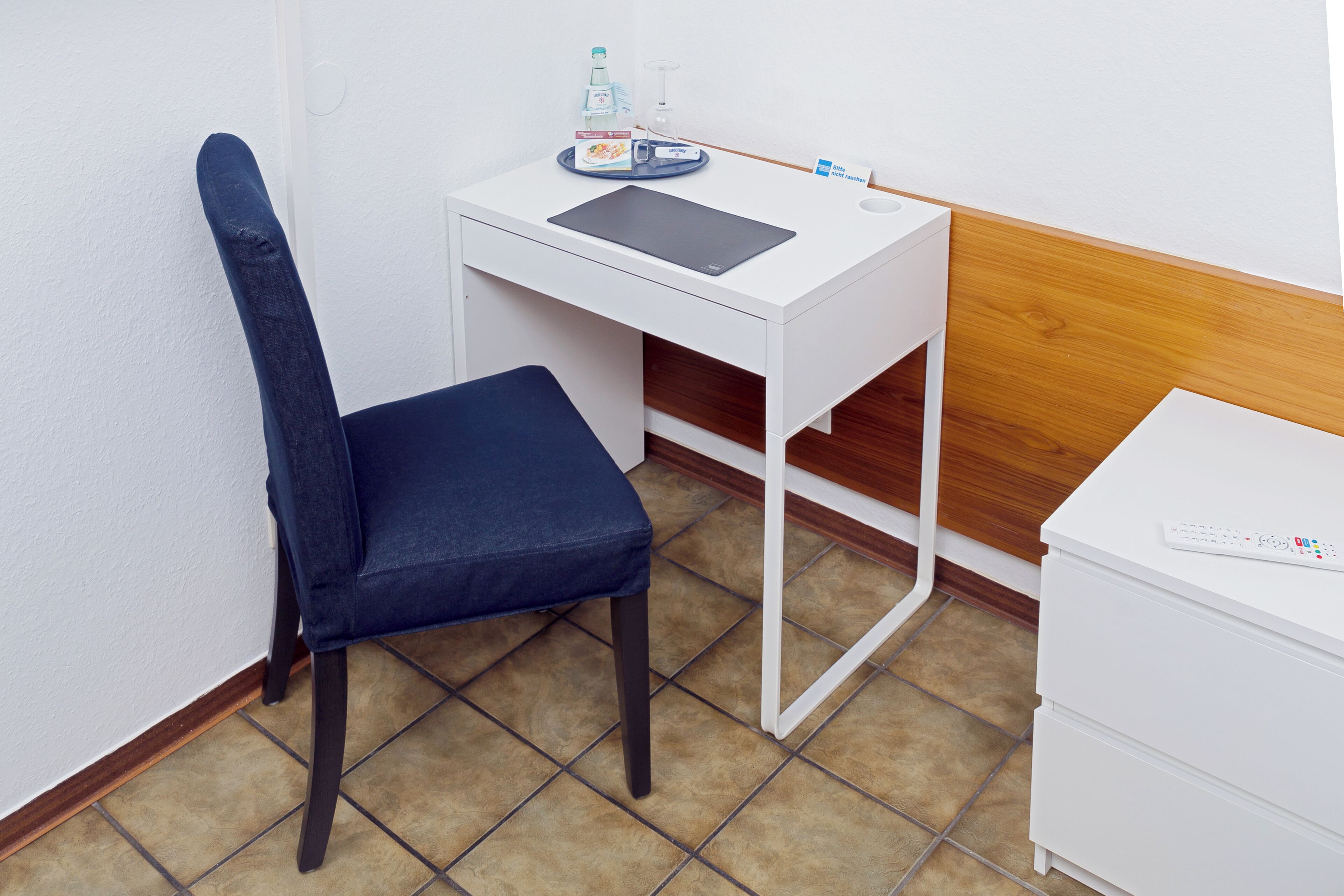 A small, white desk with a tablet, a bottle, and a glass. A blue chair is positioned in front of the desk.
