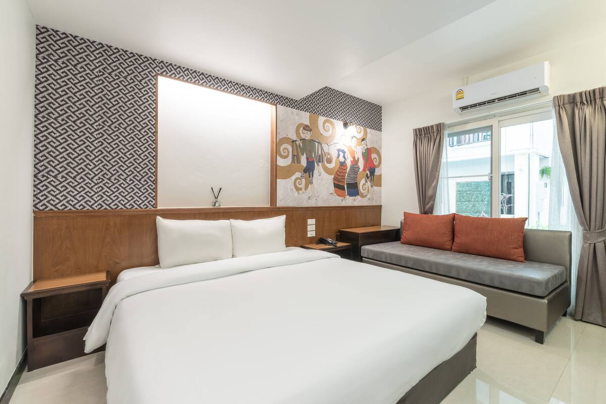 A hotel room with a large bed, a sofa, a wall-mounted air conditioner, and a patterned wall.