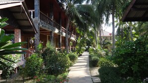 A corridor with a checkered floor, surrounded by lush greenery and palm trees, leading to a building with a balcony and hanging plants. - Family House Resort (Ko Pha-ngan)