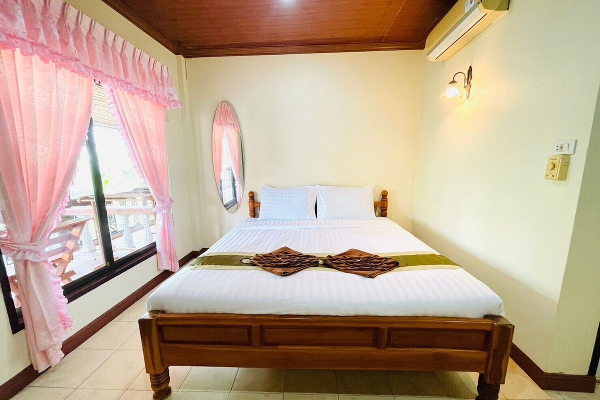 Cama bem arrumada com estrutura de madeira, edredão branco e duas almofadas decorativas. Há uma janela com cortinas cor de rosa e vista para uma varanda externa.