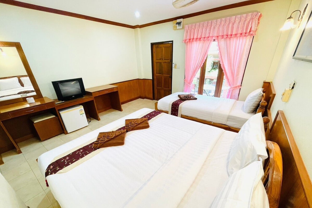 Quarto de hotel com duas camas, uma mesa de trabalho com televisão, frigorífico e janela com cortinas cor-de-rosa.