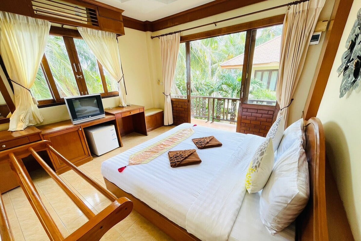 Quarto de hotel com cama, escrivaninha com televisão, mini-geladeira e varanda com vista para o verde.