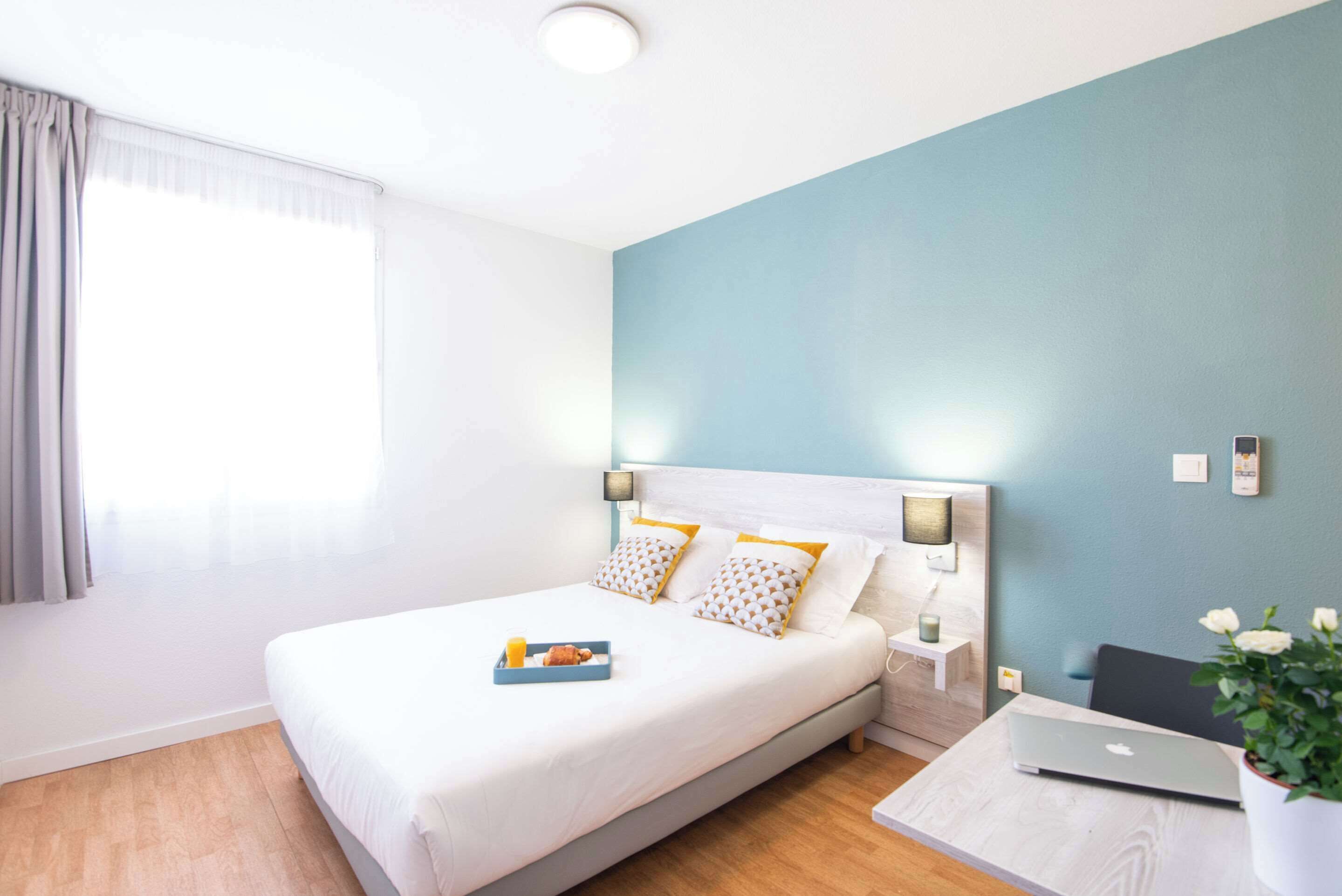 Une chambre d’hôtel avec un lit, une table de chevet avec une lampe, un ordinateur portable et une plante en pot.