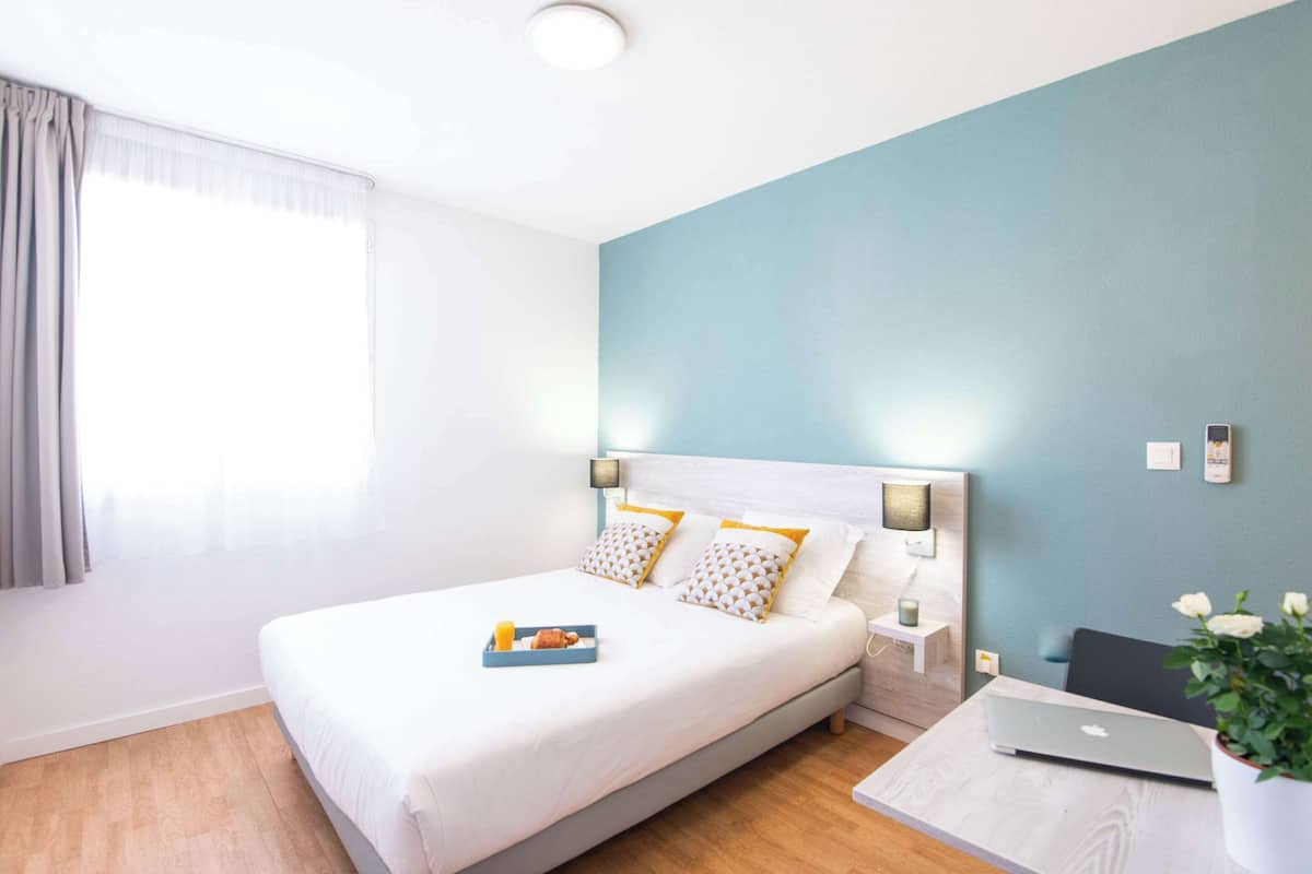 A hotel room with a bed, a bedside table with a lamp, a laptop, and a potted plant.