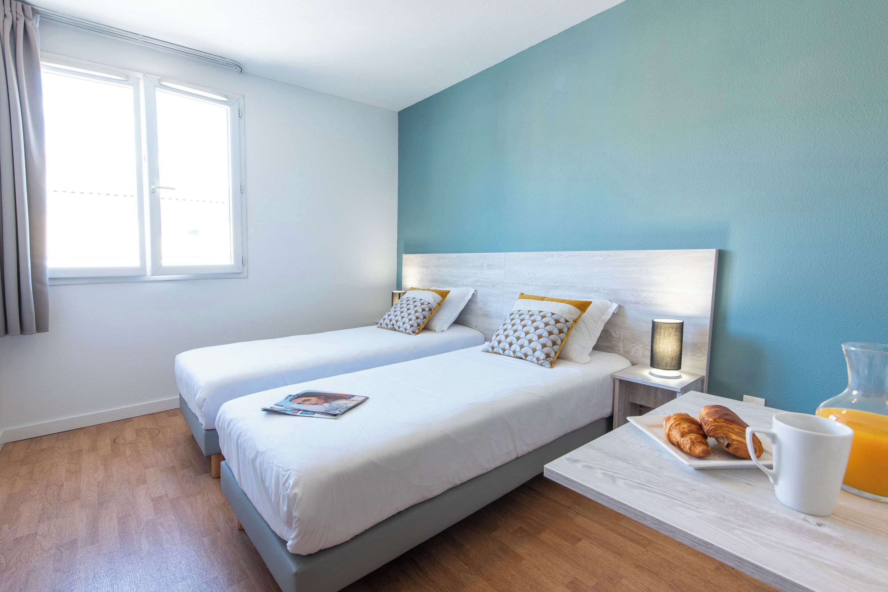 Une chambre d’hôtel avec deux lits, une table de chevet avec une lampe et une table pour le petit-déjeuner avec des croissants et du jus d’orange.