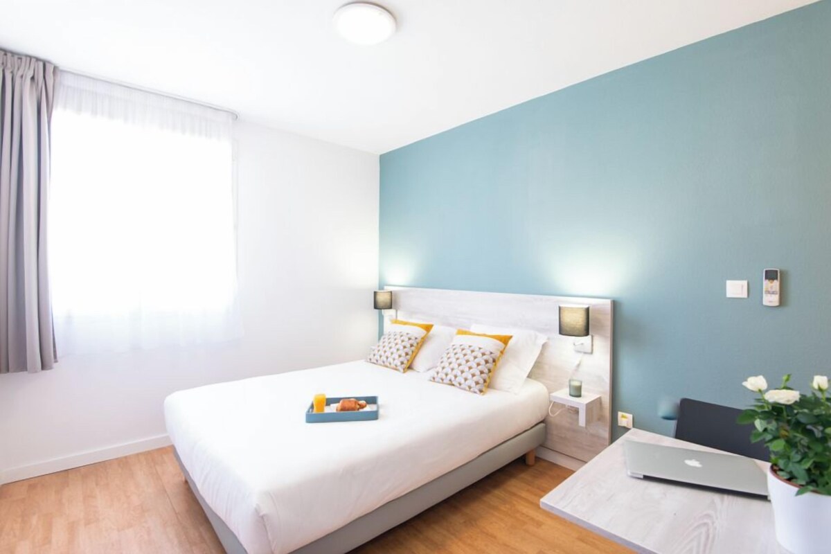 Une chambre d’hôtel moderne avec un lit, une table de chevet et un ordinateur portable posé sur un bureau.