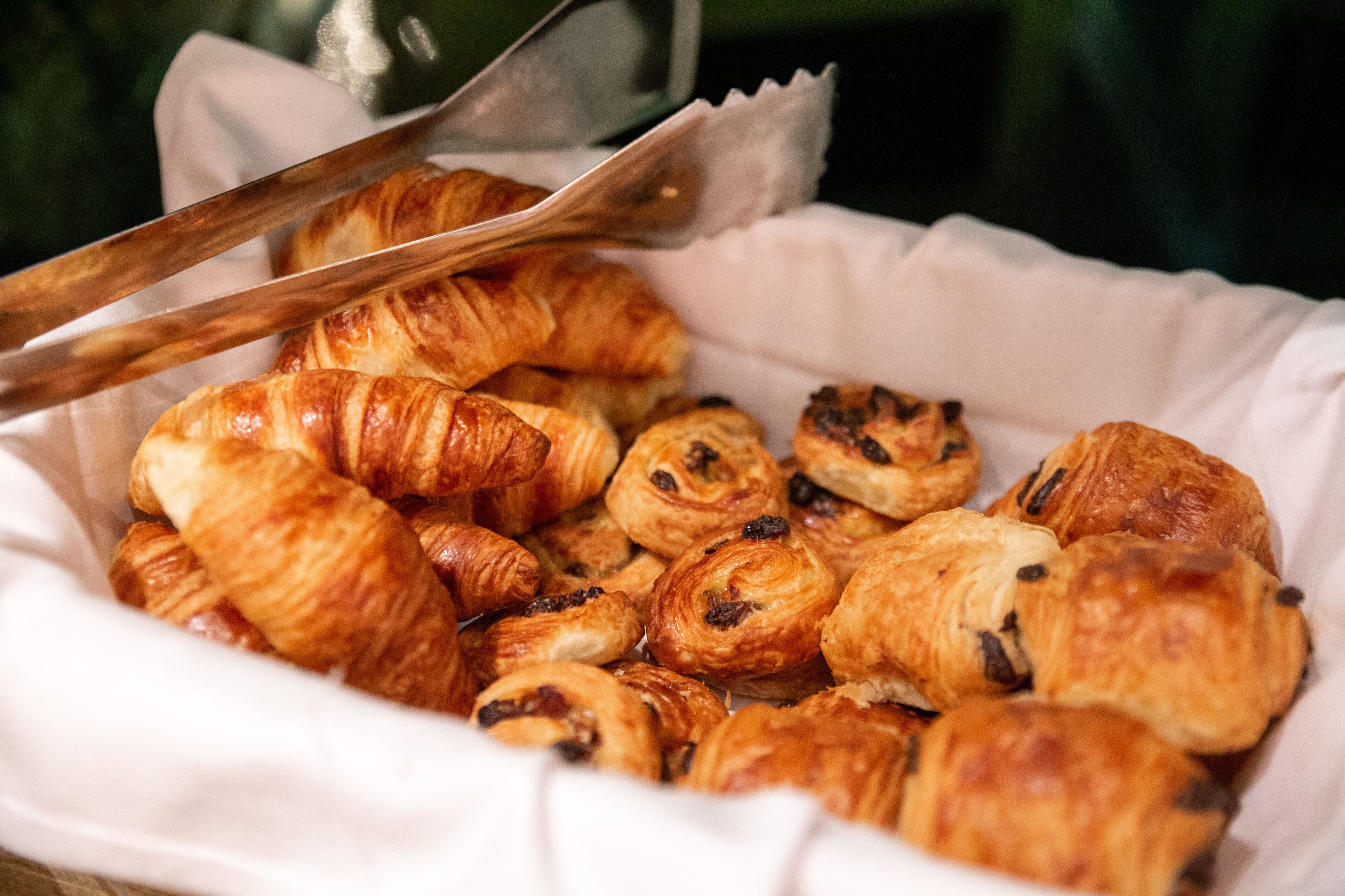 A basket of assorted pastries, including croissants and chocolate-filled pastries, with a pair of tongs resting on top.