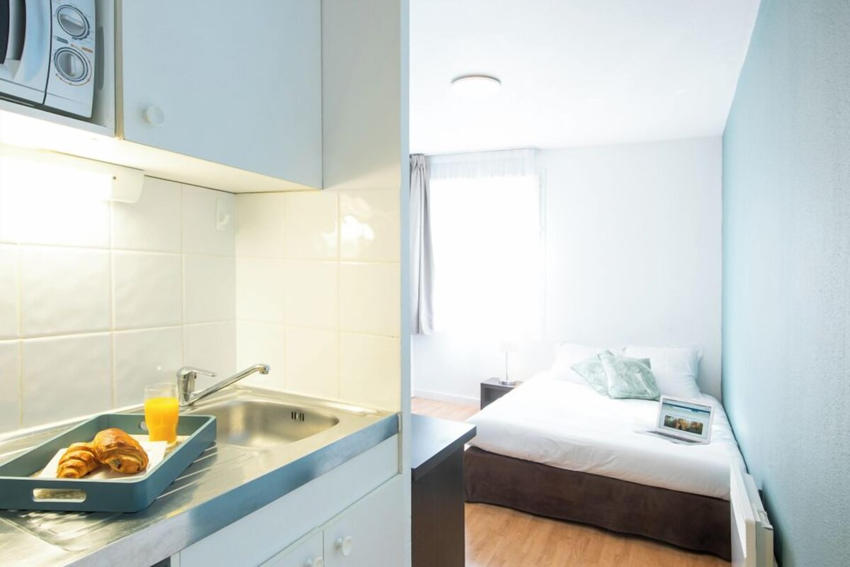 A kitchenette with a sink, a tray with croissants and orange juice, and a bed with a white comforter and pillows.