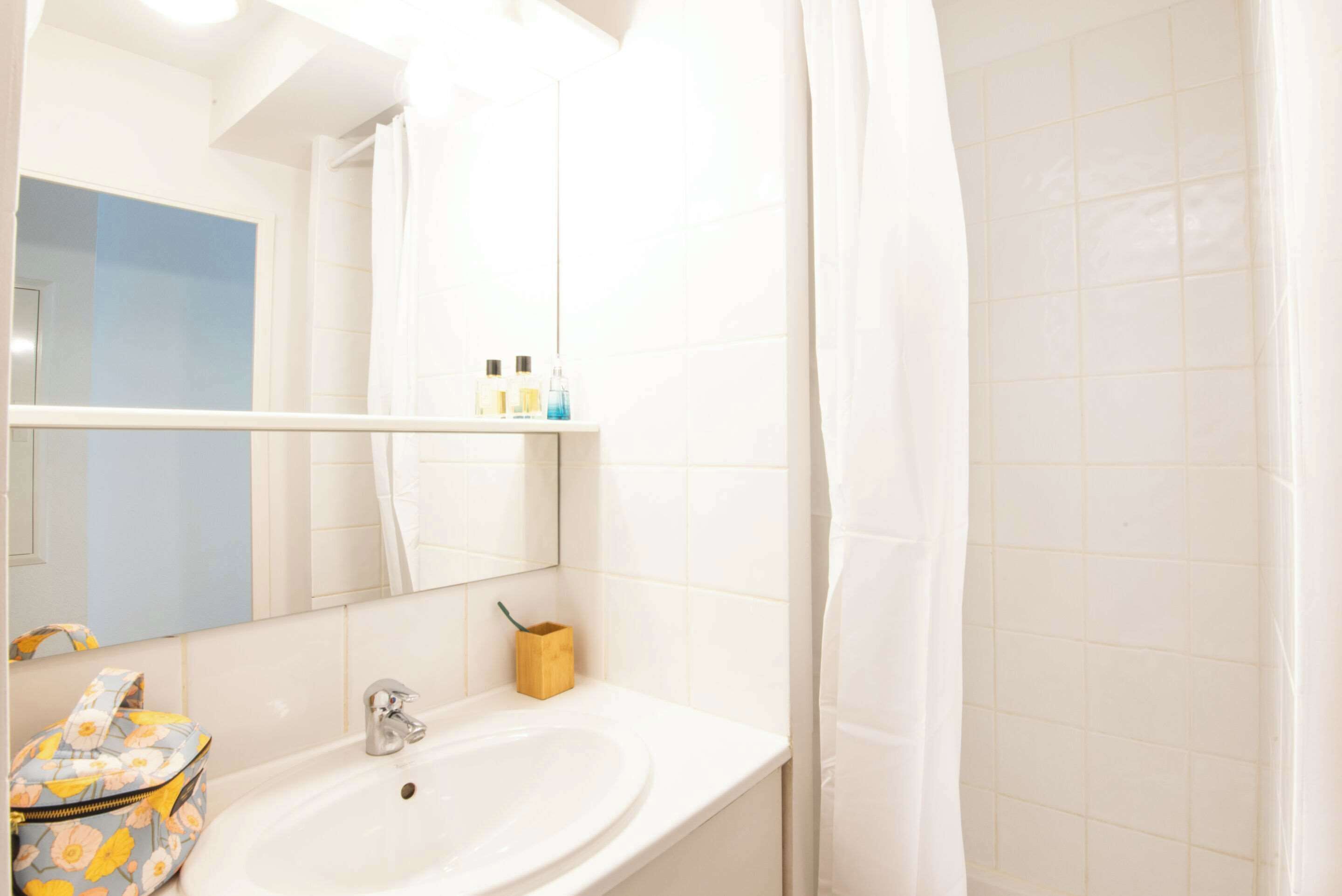 A bathroom with a white sink, mirror, and tiled walls. There is a shower curtain, a small wooden box, and a floral bag on the counter.