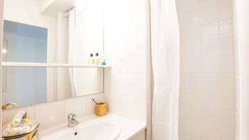 A bathroom with a white sink, mirror, and tiled walls. There is a shower curtain, a small wooden box, and a floral bag on the counter.