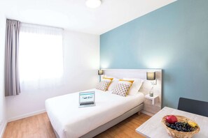 A hotel room with a bed, a laptop on the bed, a nightstand with a lamp, a fruit basket on a table, and a window with curtains.