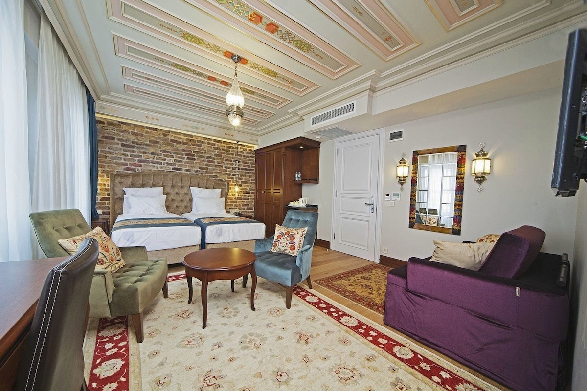 A hotel room with a brick wall, a double bed, a sofa, a chair, a small table, and a decorative mirror.
