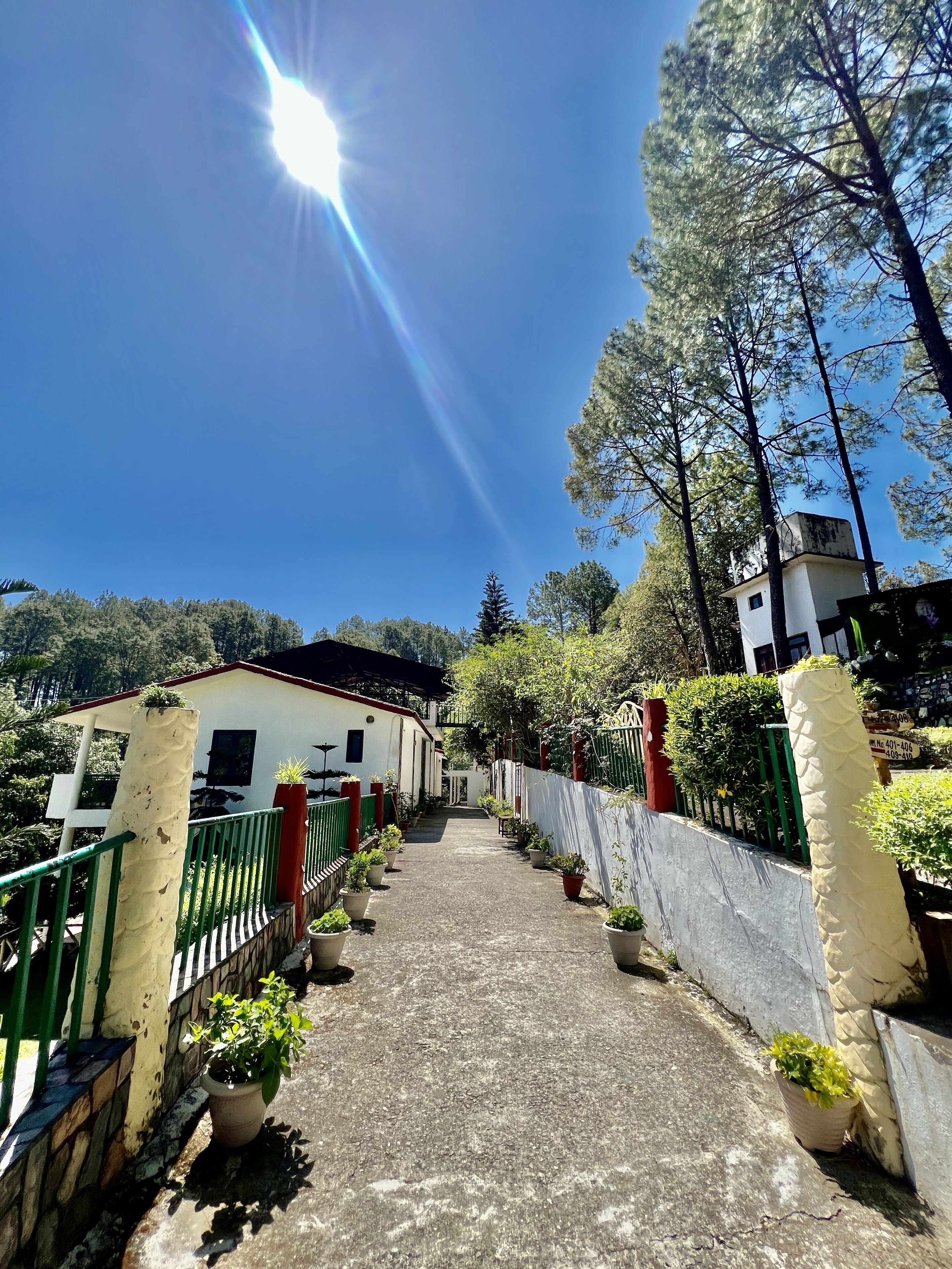 Uma trilha leva a um edifício com fachada branca e telhado vermelho, cercado por vegetação e árvores.