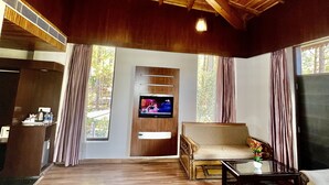 A spacious living area with a wooden ceiling, a flat-screen TV mounted on a wooden panel, a sofa, a coffee table, and a view of the outdoors through large windows. - Sea Hawk Hill Resort - A Luxury Boutique Resort (Nainital)