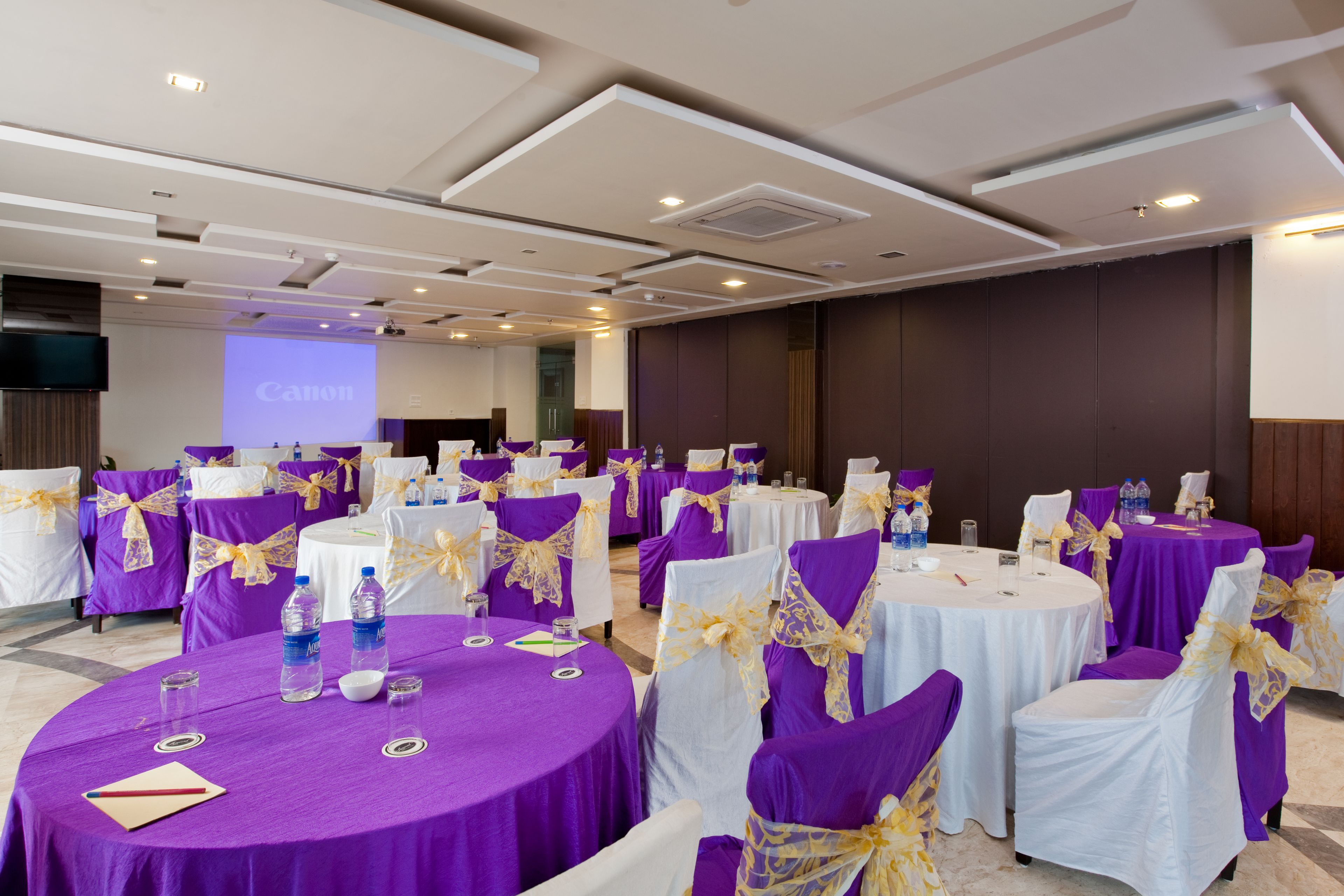 Un salón de conferencias con mesas redondas, manteles morados y blancos, y sillas con cintas amarillas.