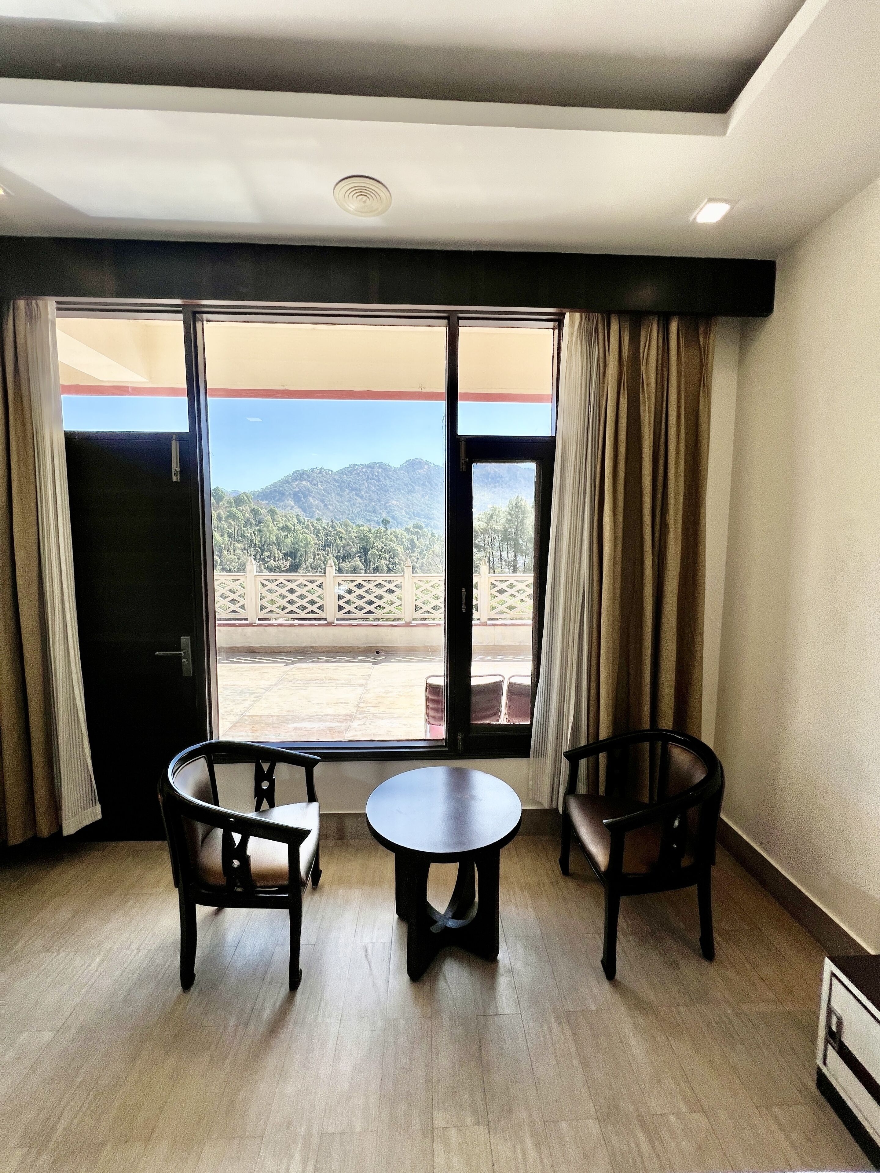 Una habitación con un gran ventanal corredizo que da a un balcón con vistas a montañas. Hay dos sillas y una mesita en la habitación.