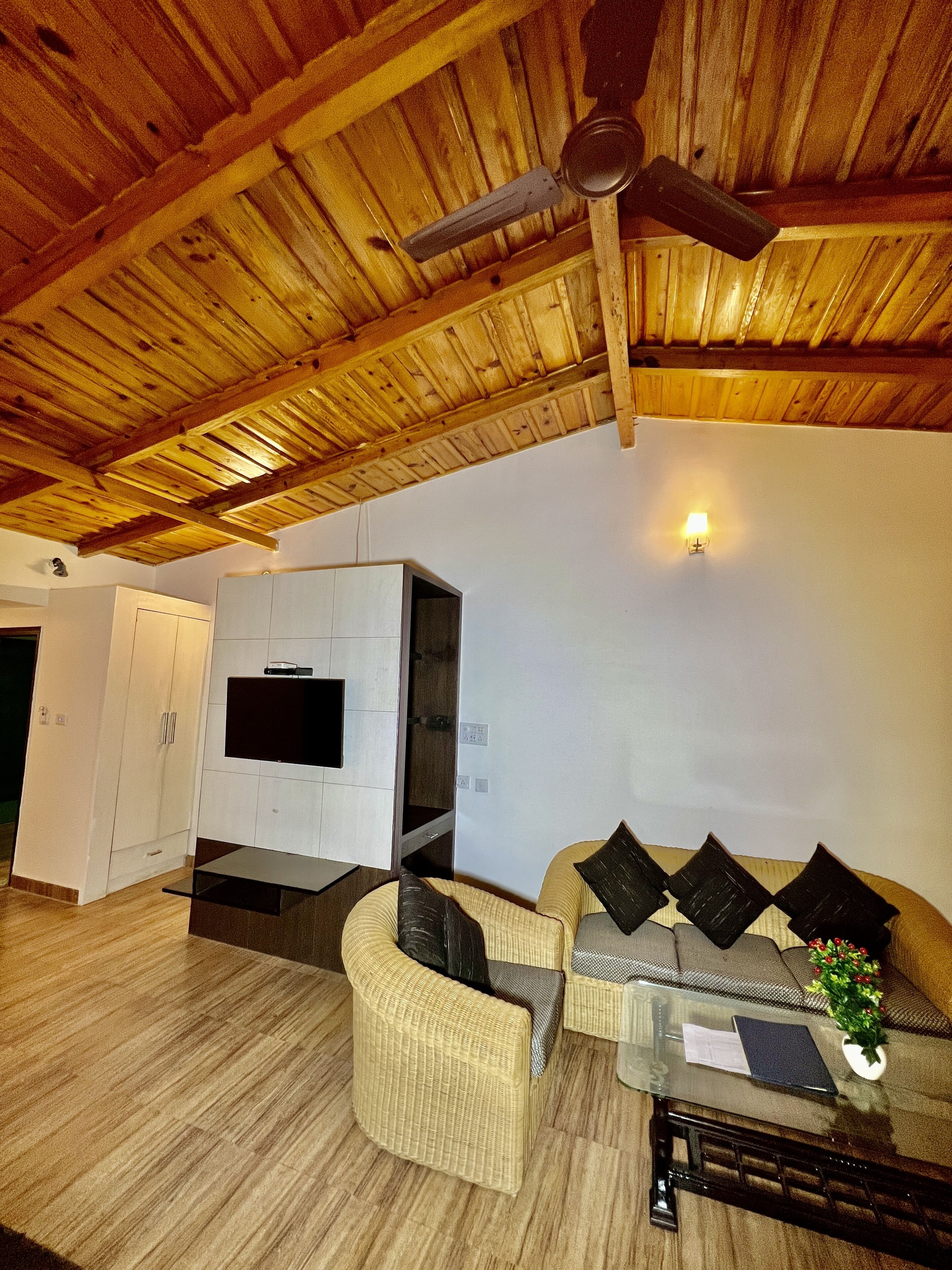 A room with a wooden ceiling, a television mounted on a wall, a sofa with cushions, a small table with a plant, and a ceiling fan.