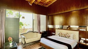 A spacious bedroom with a large bed, wooden ceiling beams, a small sitting area with a sofa, and a view of greenery outside. - Sea Hawk Hill Resort - A Luxury Boutique Resort (Nainital)
