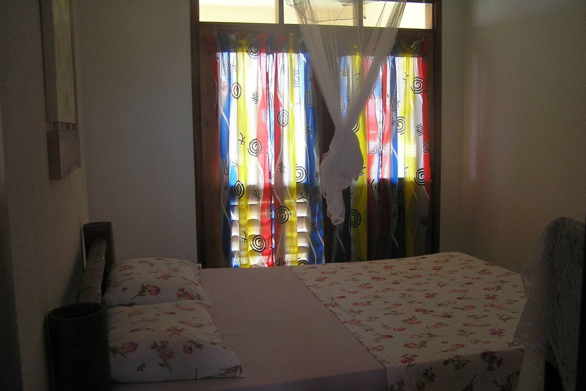 A room with two beds, a window with colorful curtains, and a mosquito net.