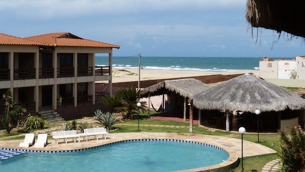 Un espace piscine avec vue sur la plage et l’océan, un bâtiment avec un toit rouge et des parasols en chaume.
