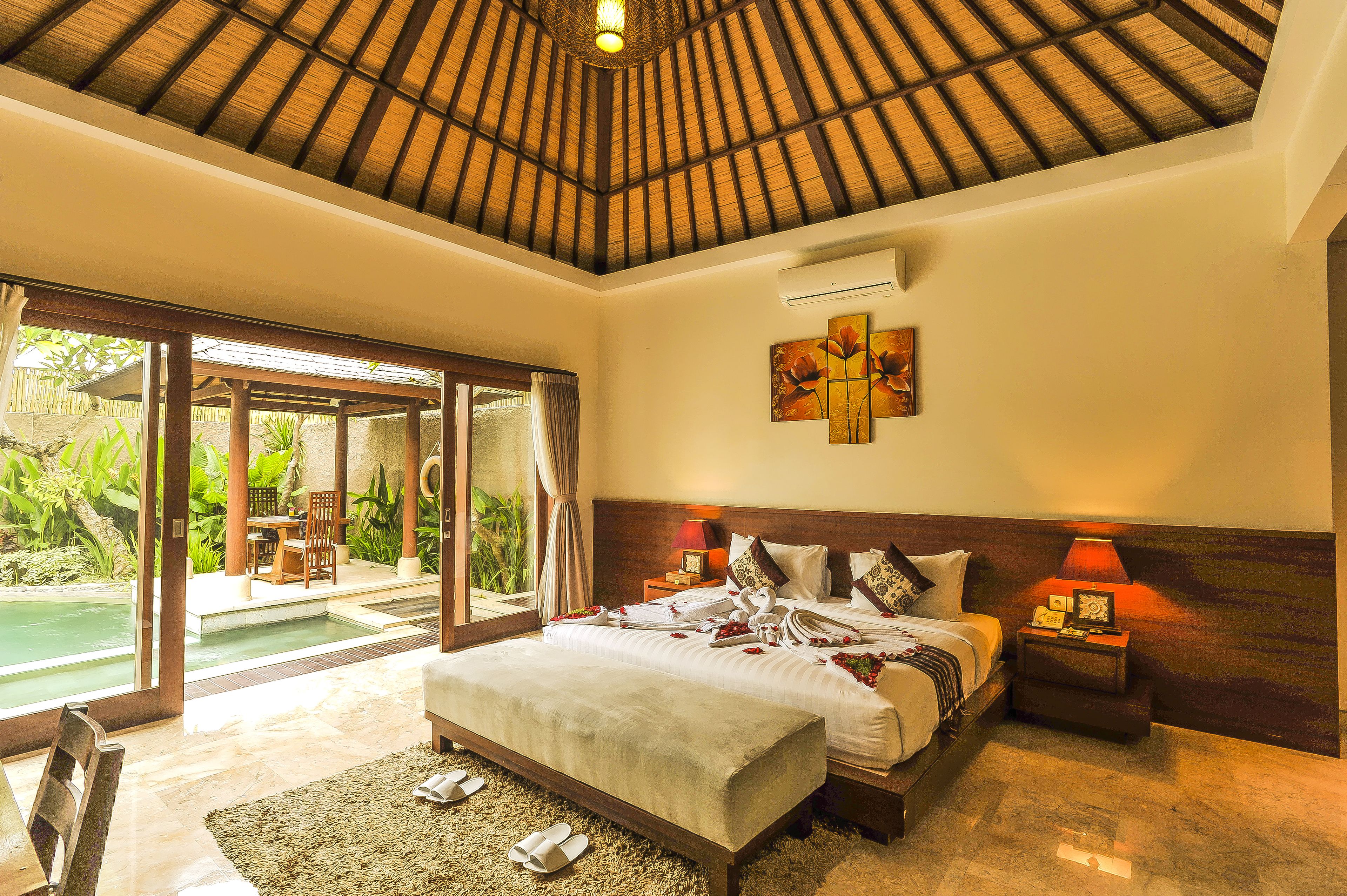 A bedroom with a large bed, a nightstand with a lamp, and a view of a pool.