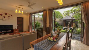 A modern living room with a flat-screen TV, sofa, dining table with floral centerpiece, and large windows offering a view of a garden and pool.