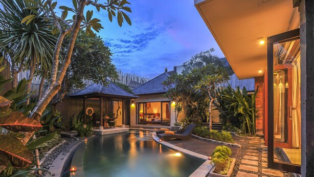 A modern outdoor pool area with a lounge chair and a small building, surrounded by tropical plants.
