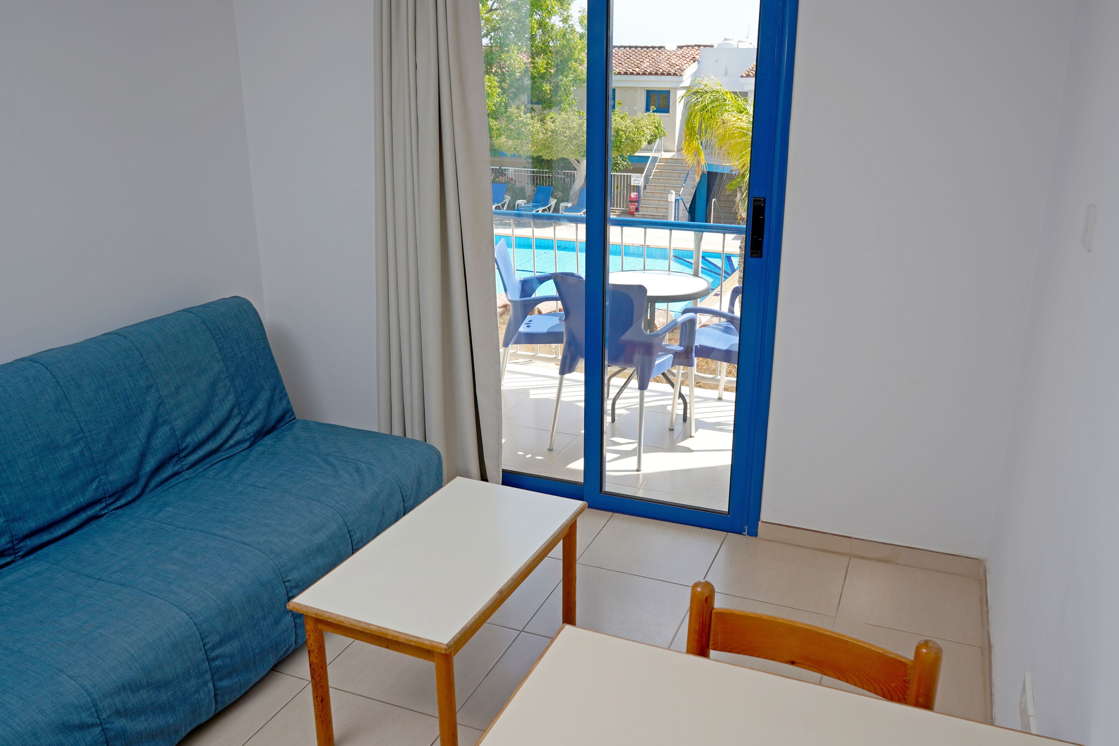 A room with a blue sofa, a small white table, and a view of an outdoor pool area through a glass door.