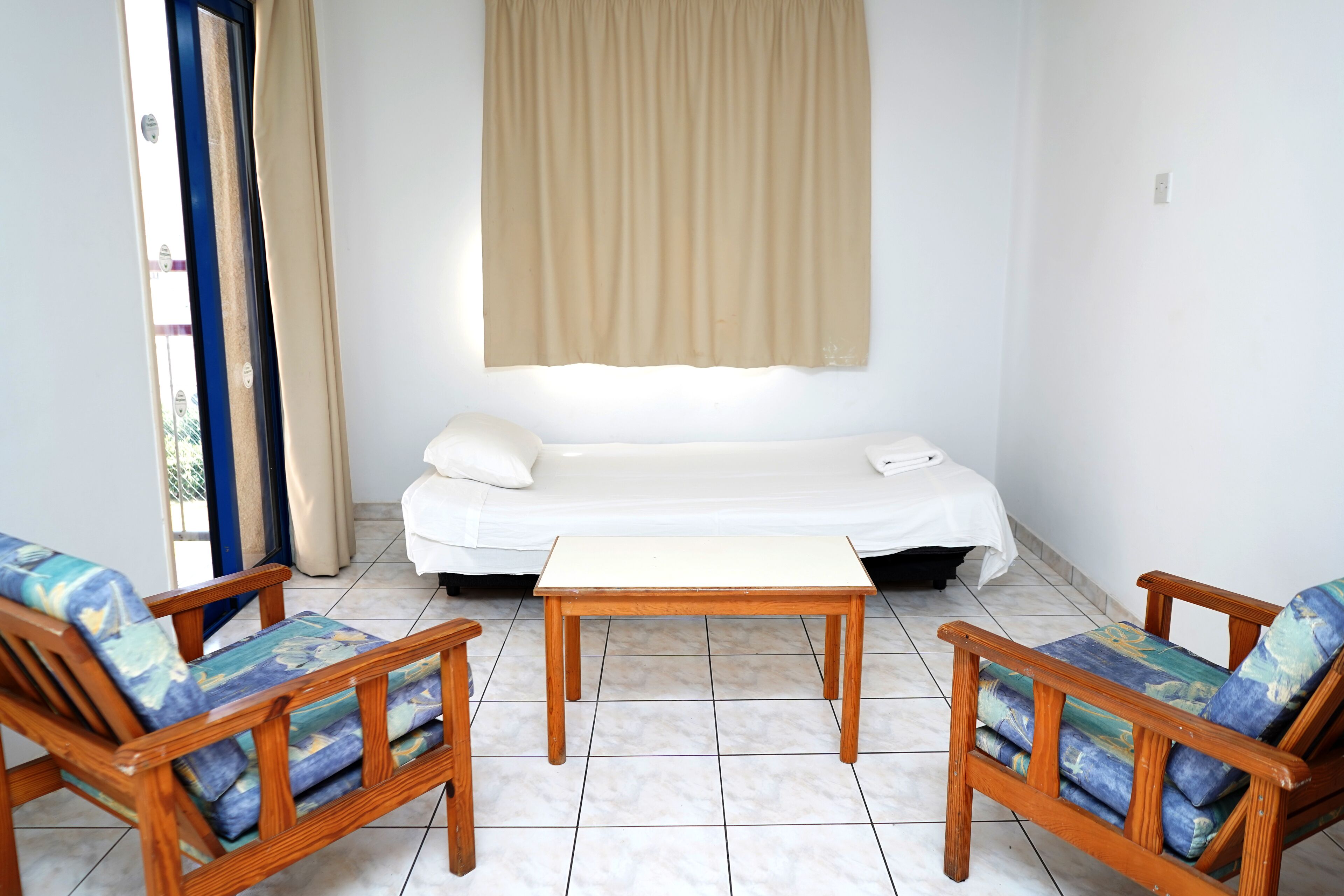 Une pièce comprenant un lit, deux chaises en bois avec des coussins, une petite table en bois et un rideau beige.