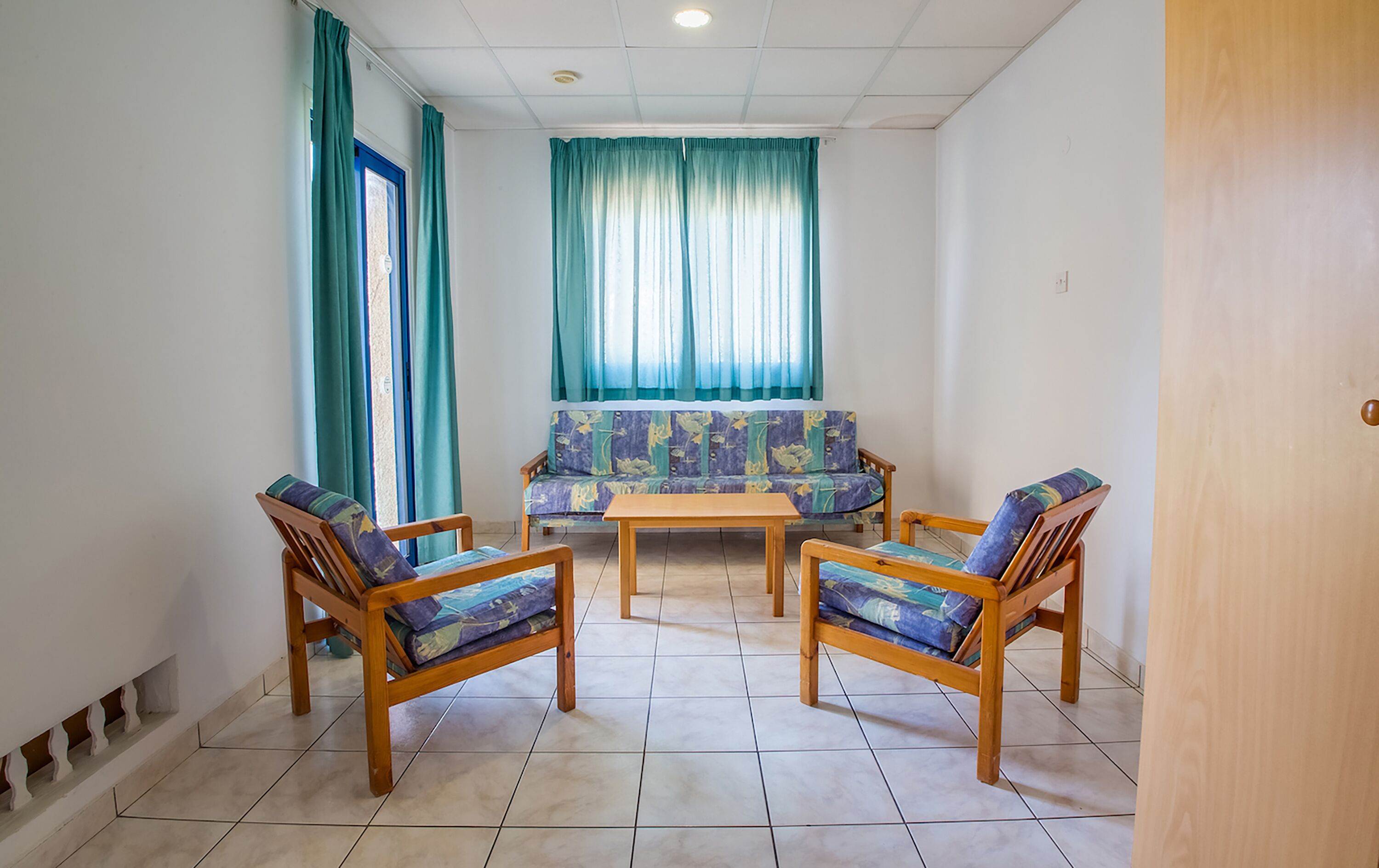 A room with a sofa set, two wooden chairs, a small table, and a door.