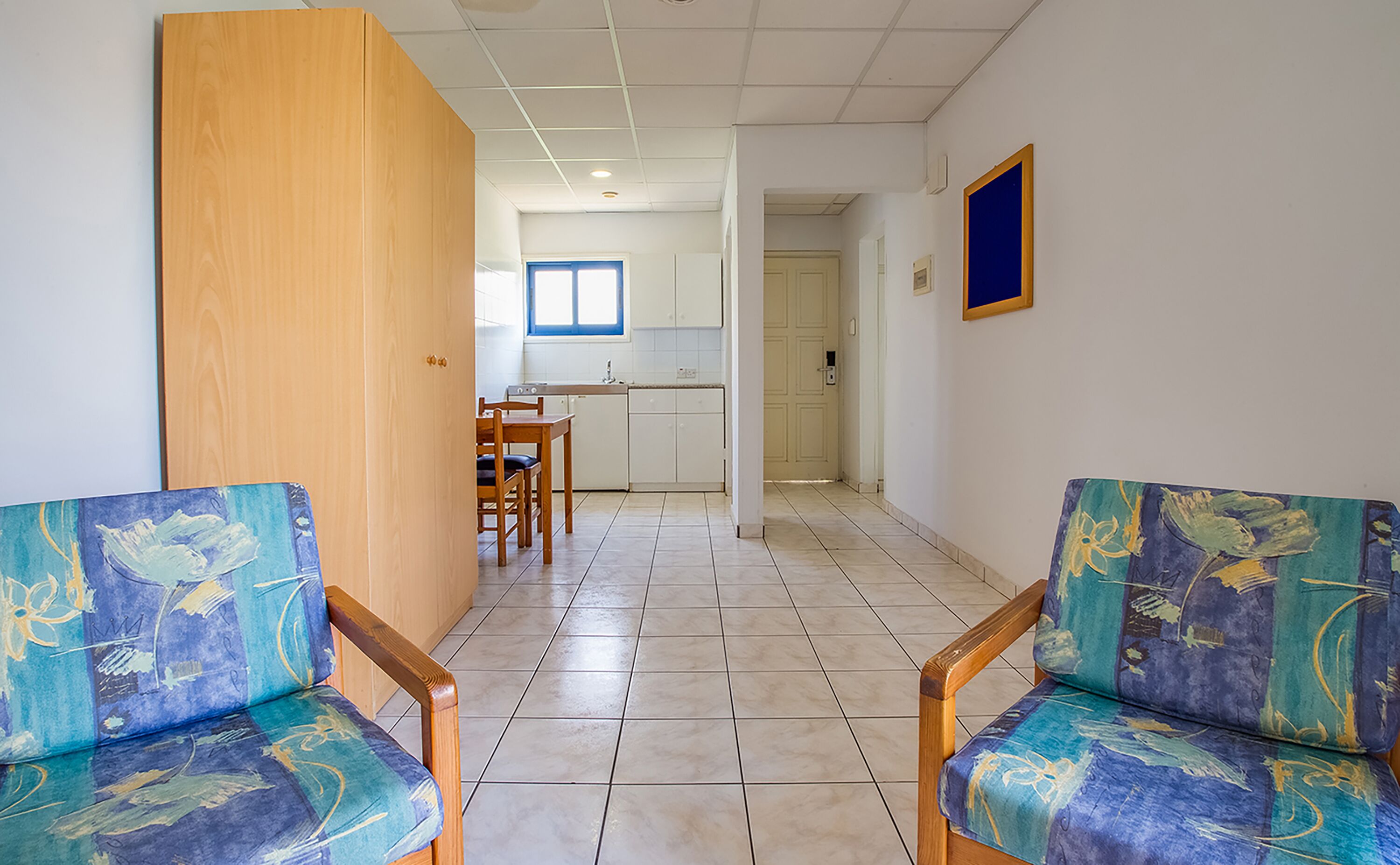 Un pasillo con dos sillas de madera con tapicería azul y verde, una puerta de madera y una zona de cocina al fondo.