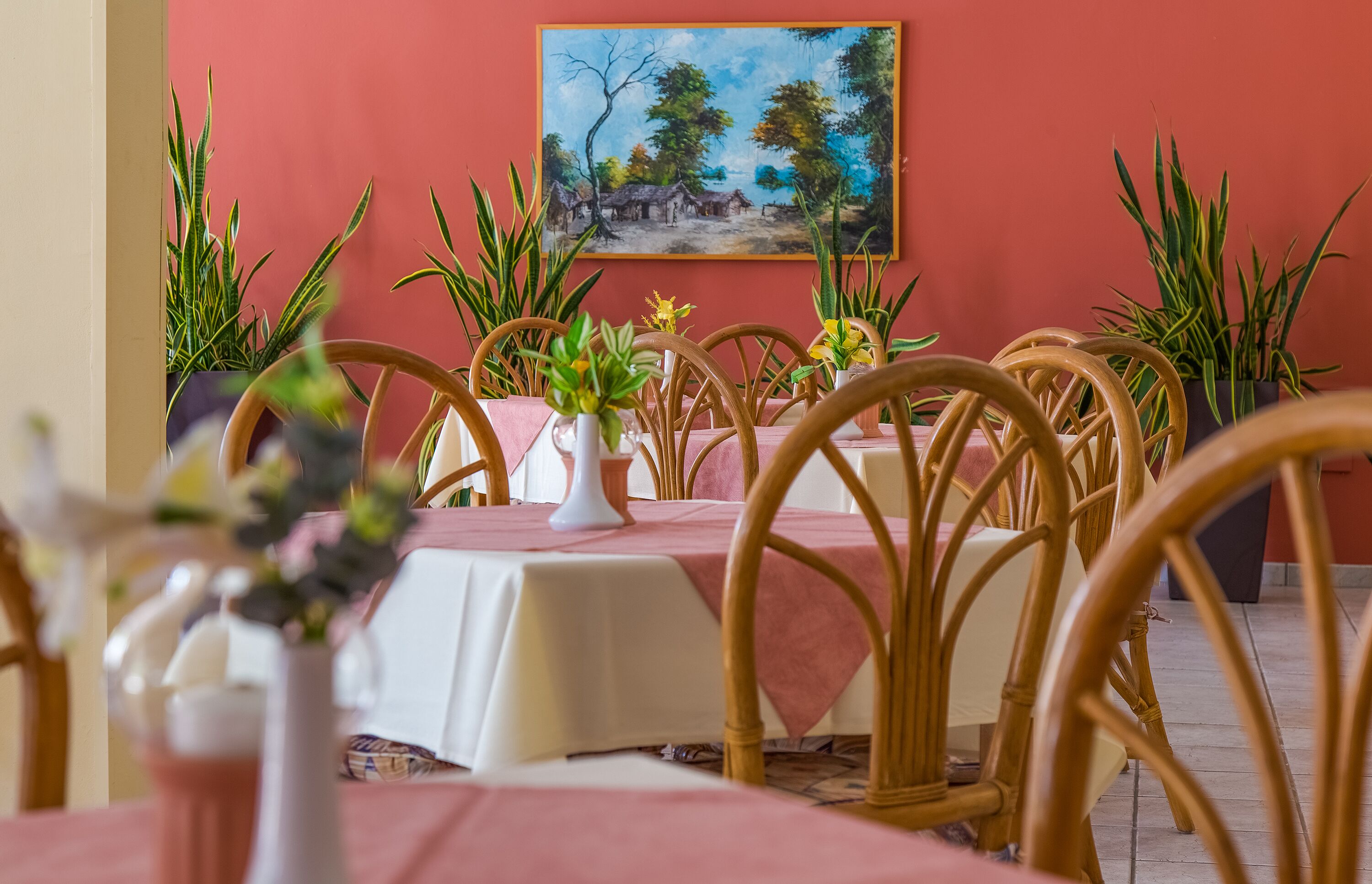 Un coin repas avec des tables préparées pour accueillir quatre personnes, entouré de chaises en osier, agrémenté de plantes en pot et orné d’un tableau au mur.